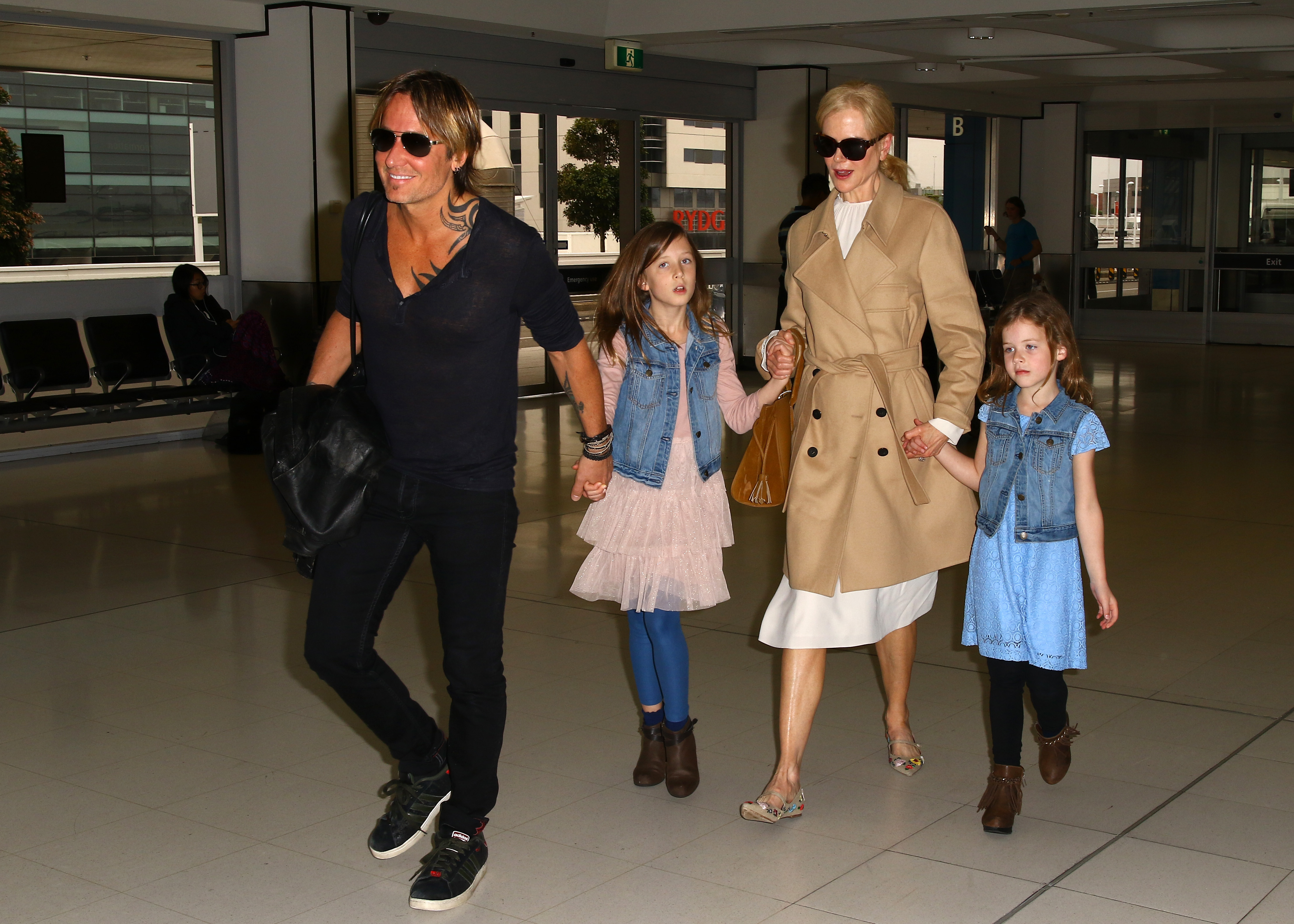 Nicole Kidman and Keith Urban walk hand in hand with their daughters, Sunday Rose and Faith Margaret, through Sydney Airport, on March 28, 2017. The couple appears coordinated in neutral tones, with Nicole in a beige trench coat and sunglasses, while both girls wear denim vests over pastel dresses as the family makes their way through the terminal.