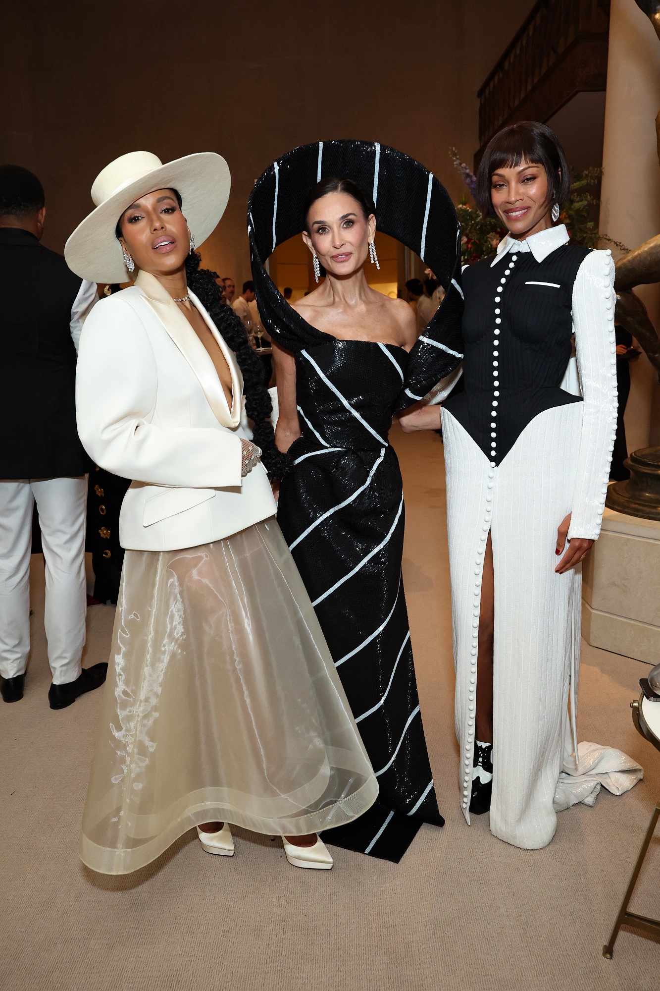 Moore stands between Kerry Washington, who wears a sheer ivory skirt with a plunging satin-lapel blazer and wide-brim hat, and Zoe Saldaña, who stuns in a black-and-white gown with bold buttons and a corset-inspired silhouette.