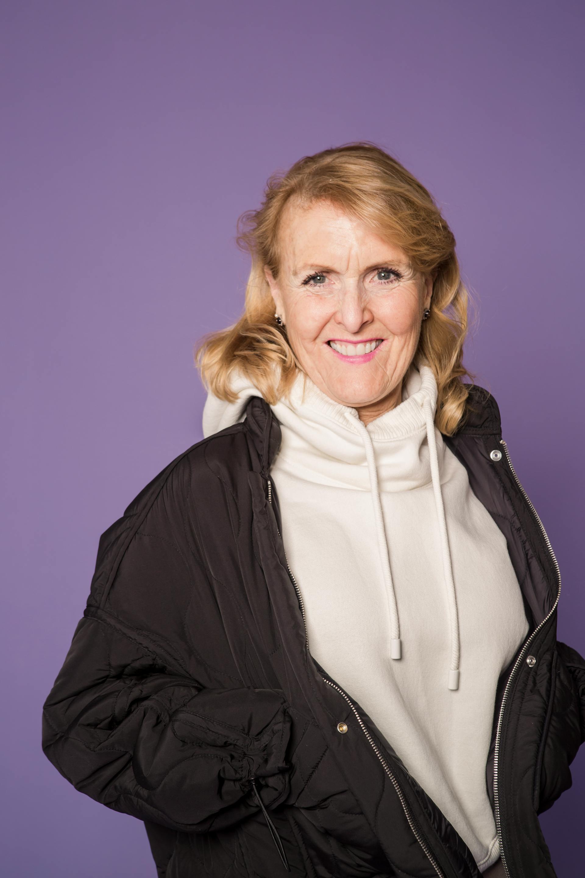 Una mujer madura y feliz con chaqueta negra y sudadera blanca sonriendo sobre un fondo morado | Fuente: Pexels