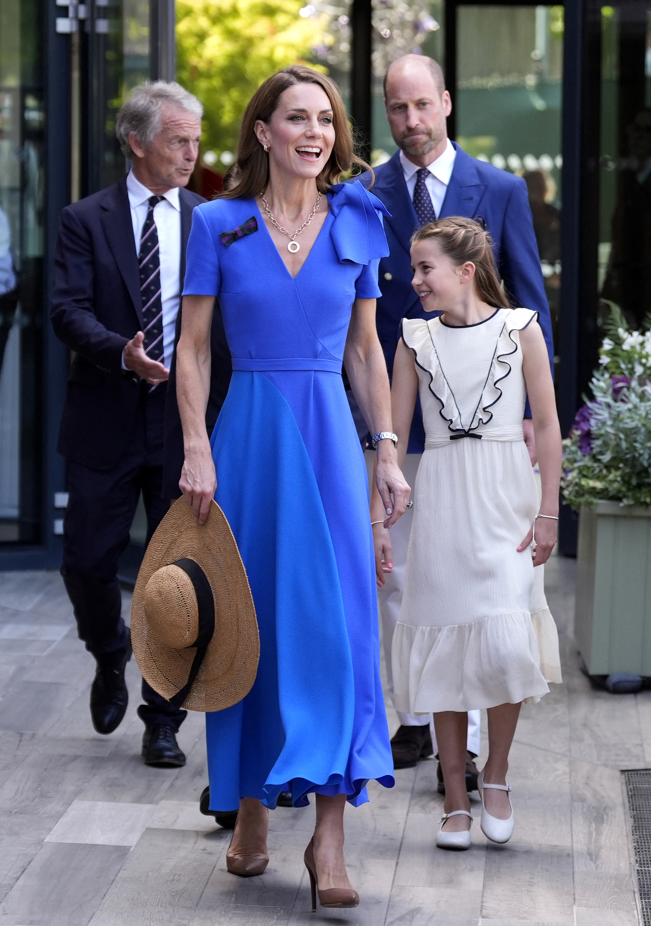 Princess Catherine walks beside Princess Charlotte while holding her sunhat in one hand. Behind them, Prince William follows as they attend the event together.