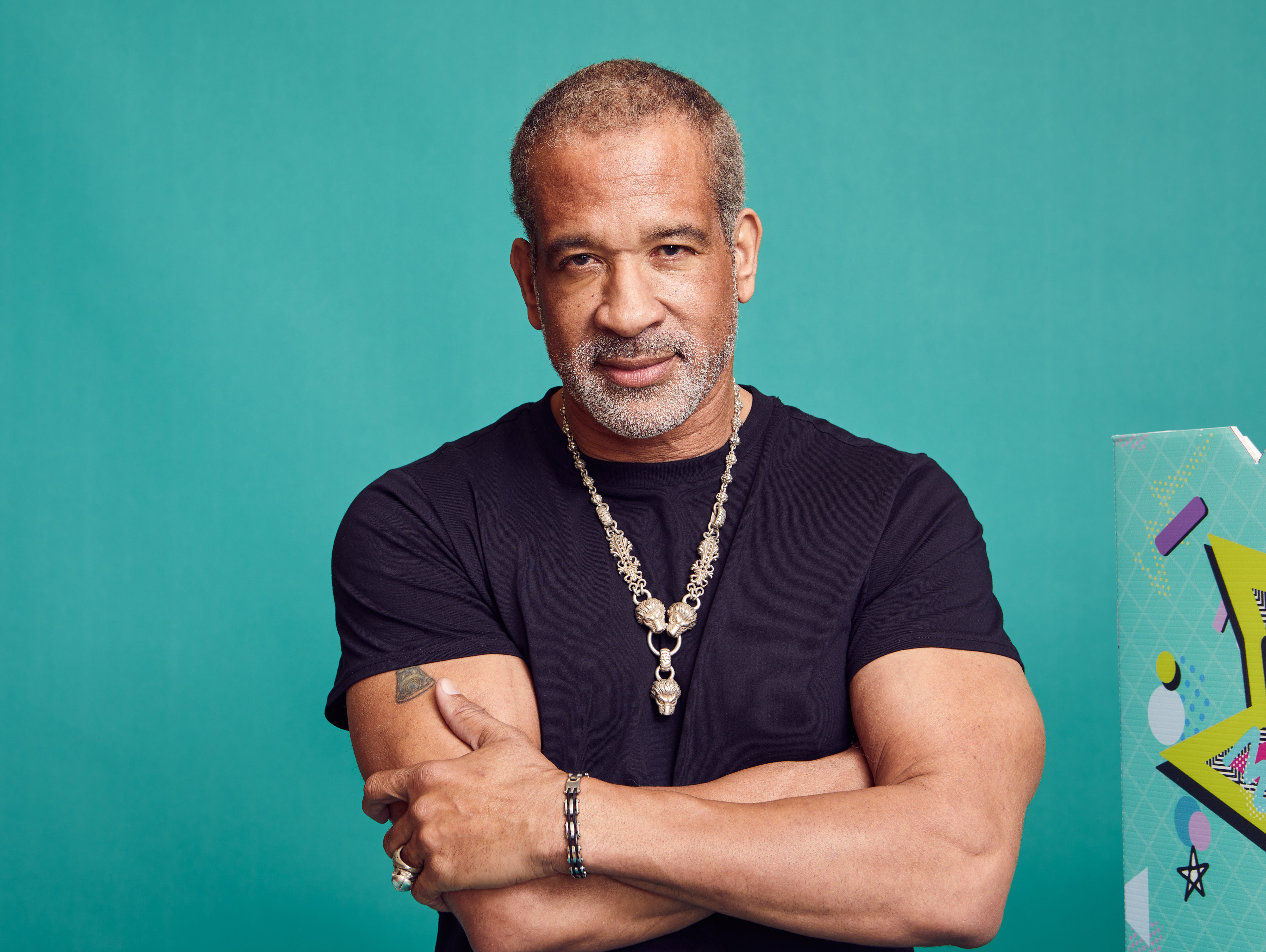 Dorian Gregory poses for a modern portrait in 2023, standing confidently against a teal backdrop with his arms crossed. Wearing a fitted black T-shirt and bold silver jewelry, he projects strength and charisma. His salt-and-pepper beard and calm, direct gaze reflect a grounded, self-assured presence.