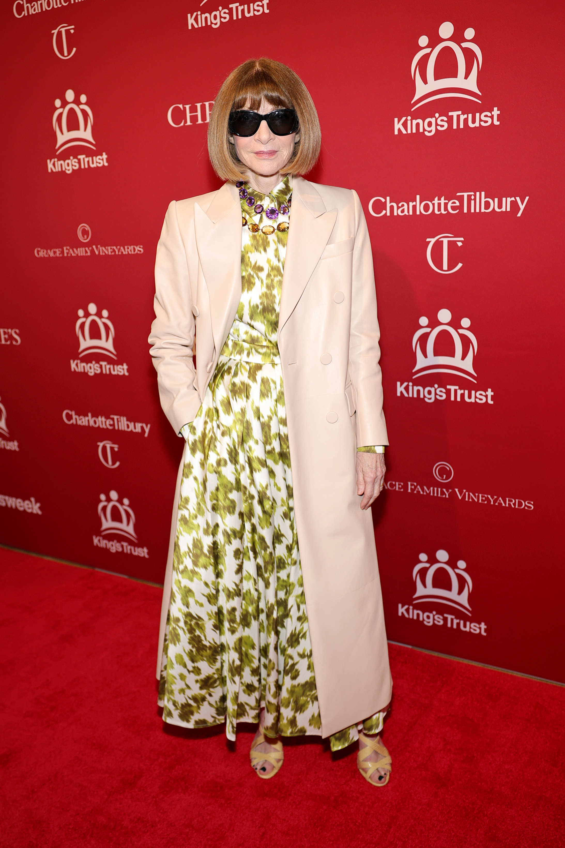 Anna Wintour poses in her full ensemble on the step-and-repeat: the olive-and-white floral dress visible beneath an open blush coat, her jewelled necklace and strappy olive sandals catching the eye, sunglasses firmly in place as she stands one hand in pocket.