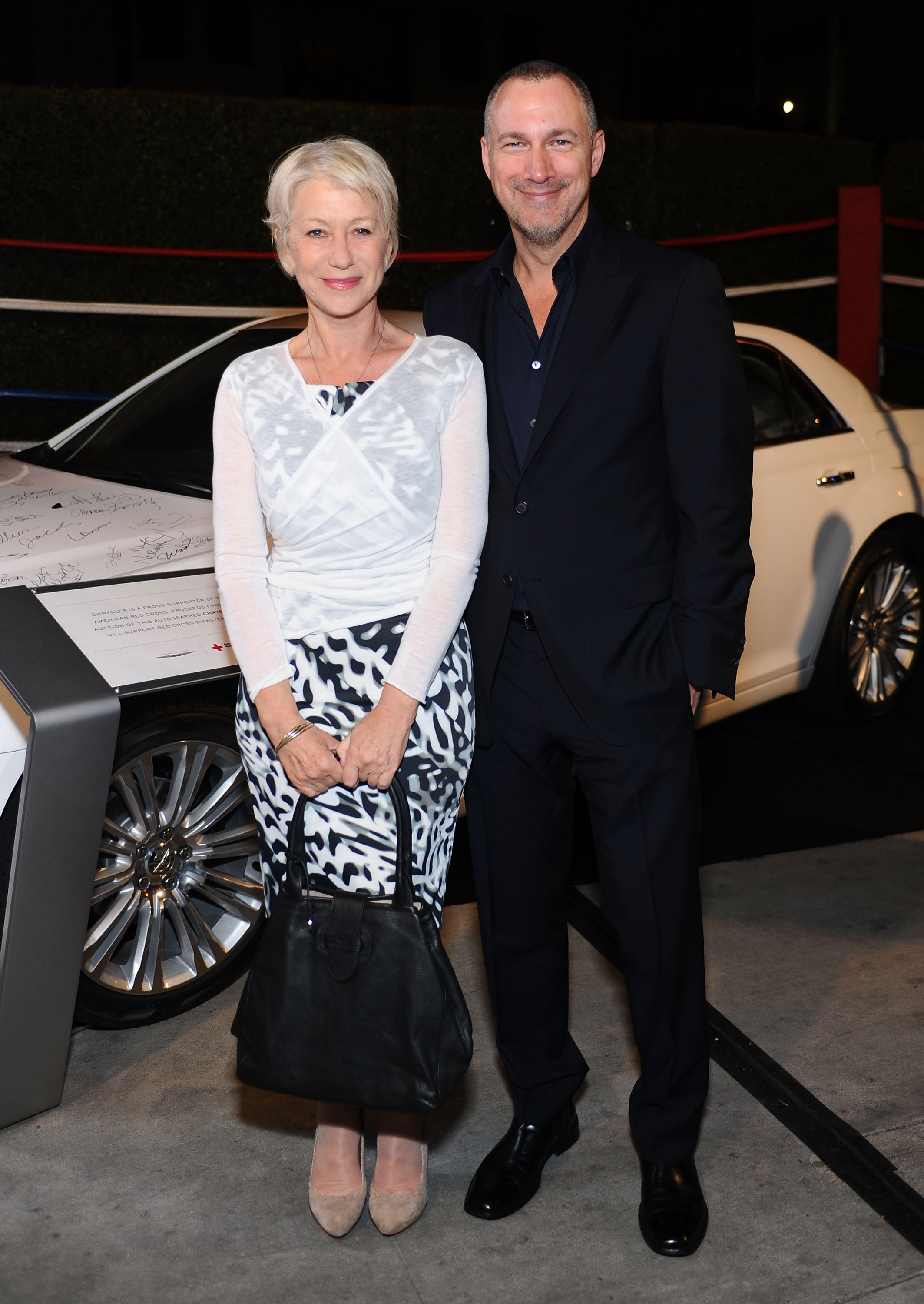 Helen Mirren poses alongside Edward Menicheschi at Vanity Fair Campaign Hollywood, wearing a chic black-and-white patterned dress layered with a sheer white wrap top, accessorized with nude heels and a classic black handbag, her short platinum hair styled elegantly.