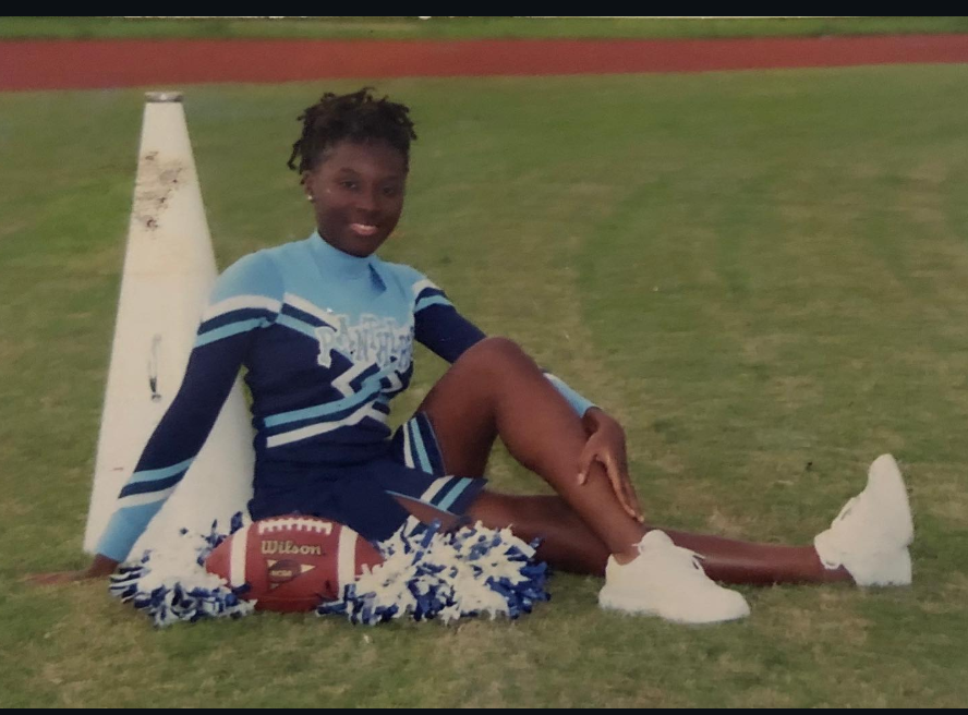 Seated confidently on the grass, a young Nancy Metayer Bowen smiles in her cheer uniform, pom-poms and a football at her side — an early glimpse of her spirited, team-driven nature. | Source: Instagram/nancymetayerbowen