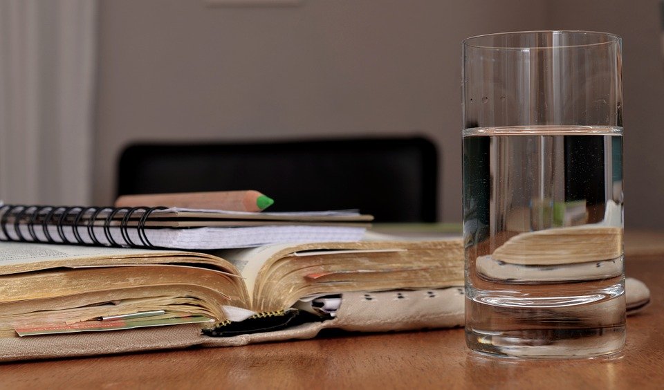 A glass of water on a table beside a book| Photo: Pixabay