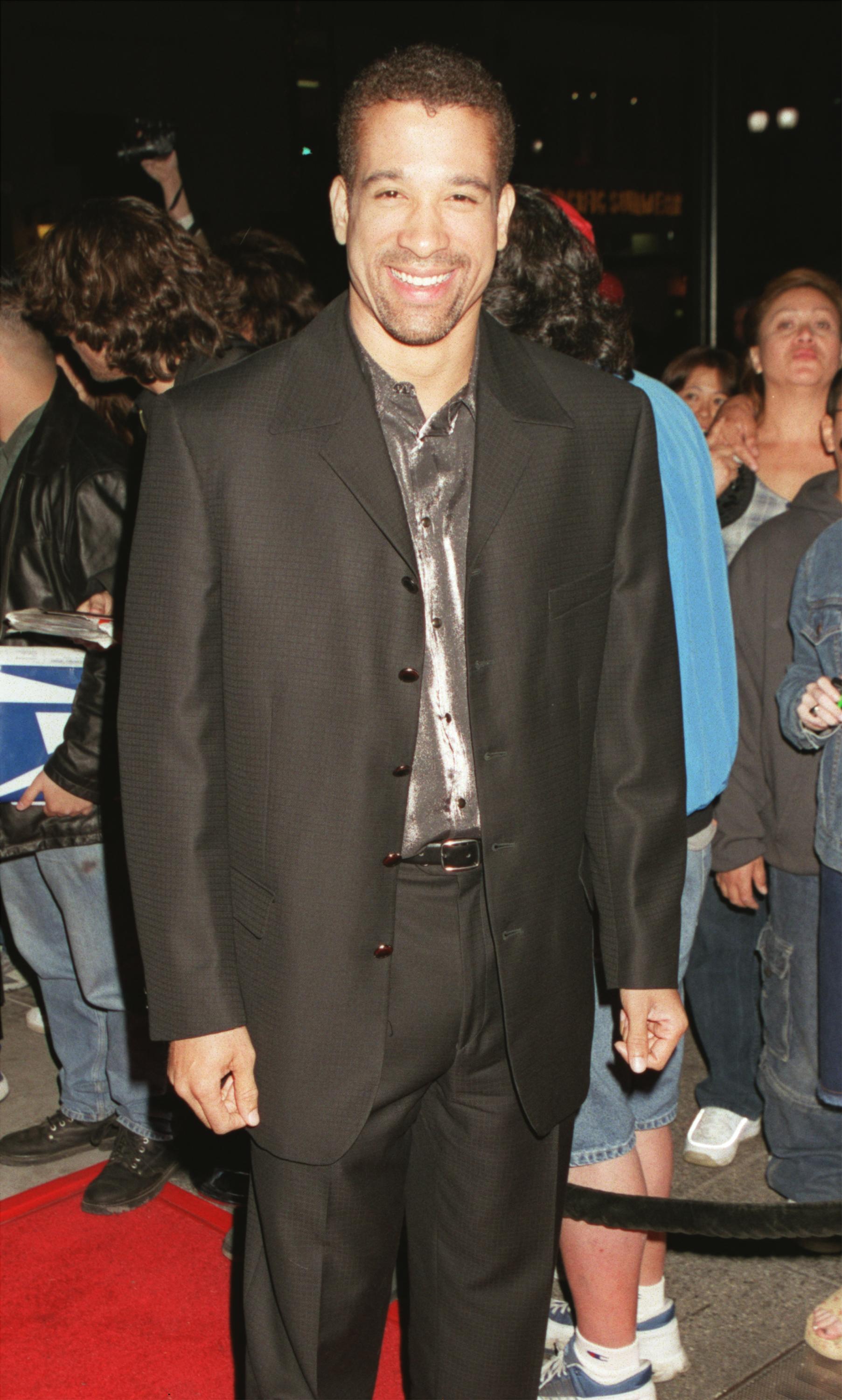 Dorian Gregory poses on the red carpet at a premiere event in the early 2000s, wearing a black suit over a shiny silver shirt. Surrounded by fans and photographers, he smiles warmly for the camera, exuding a relaxed, charismatic energy in the lively nighttime setting.