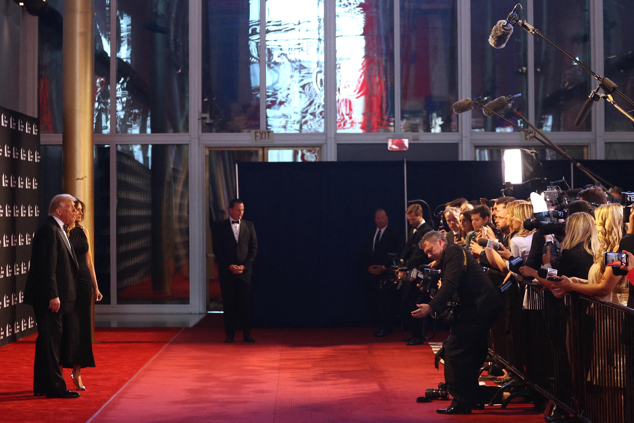 Donald and Melania stand facing a packed line of photographers, lights flashing and microphones aimed in their direction. From this wide-angle view, they appear calm and composed under the spotlight, holding their position as the press captures the moment.