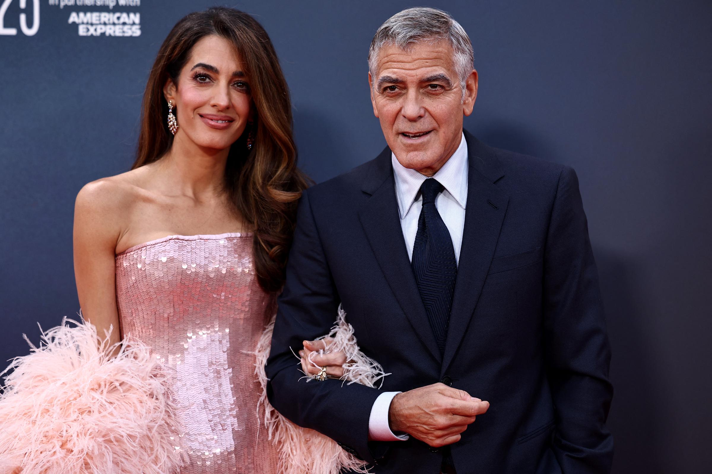 Walking arm in arm, George and Amal Clooney arrive at the BFI London Film Festival gala screening of "Jay Kelly." Amal dazzles in a strapless blush pink sequin gown with matching feathered sleeves, while George wears a dark suit with a collared shirt and tie.