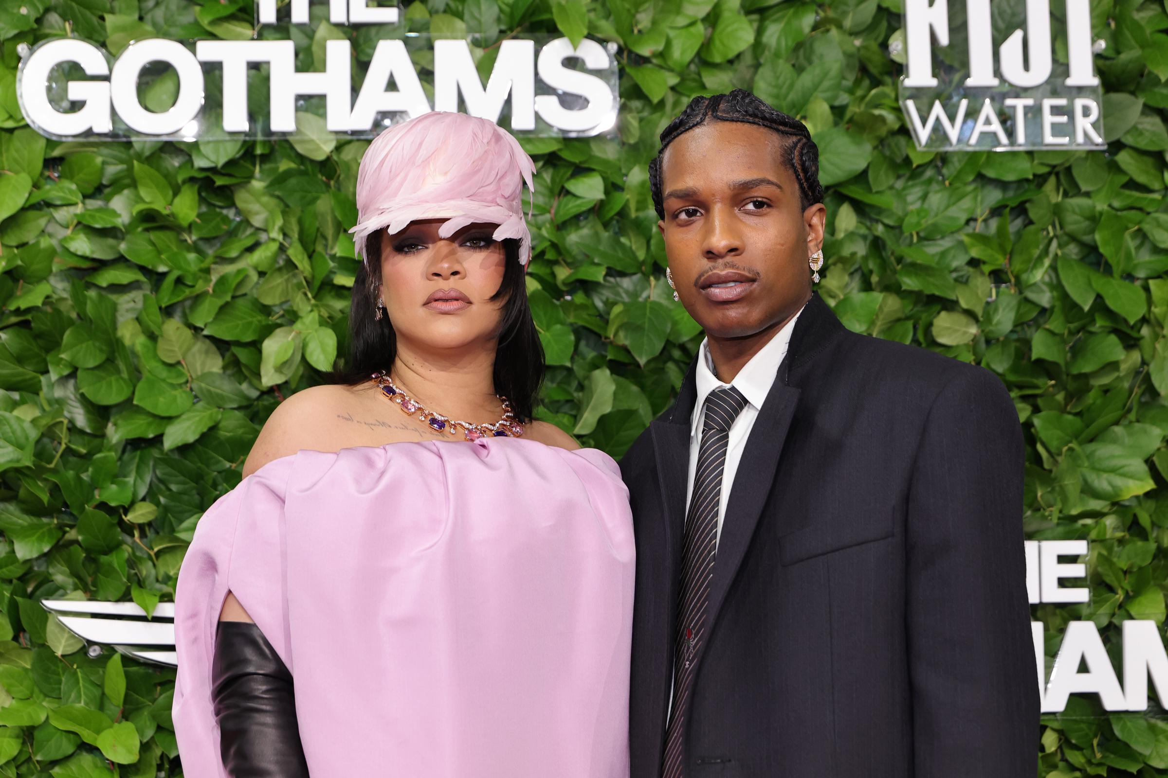 Rihanna and A$AP Rocky stand side by side on the red carpet, serving coordinated elegance — she in soft pink with gloves, he in a sleek black suit and tie, both exuding quiet confidence.