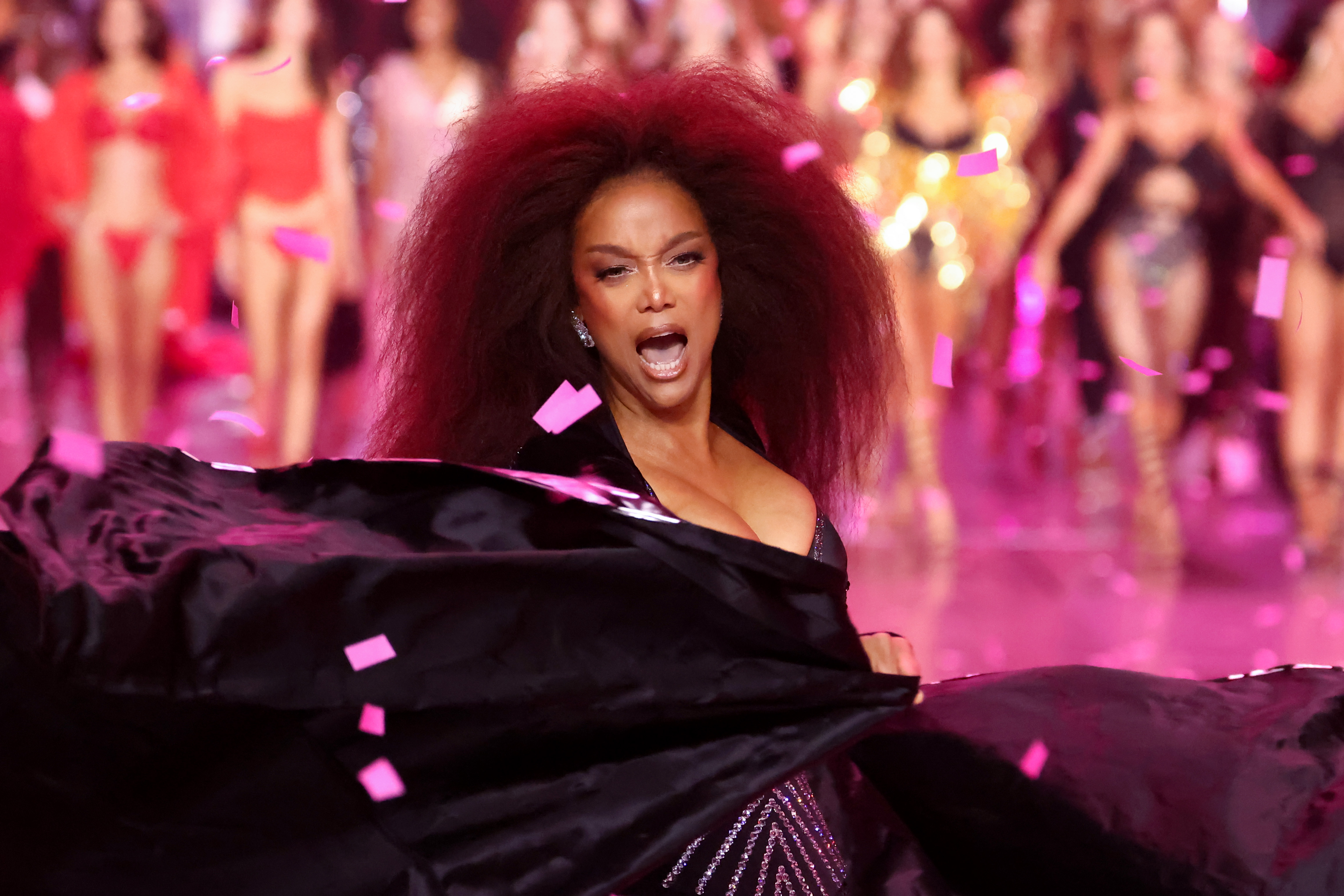 Tyra Banks unleashes high-energy glamour, whipping her dramatic cape mid-runway. Her sparkling black corset and voluminous hair heighten the moment as she shouts with fierce joy, surrounded by pink confetti and fellow models.