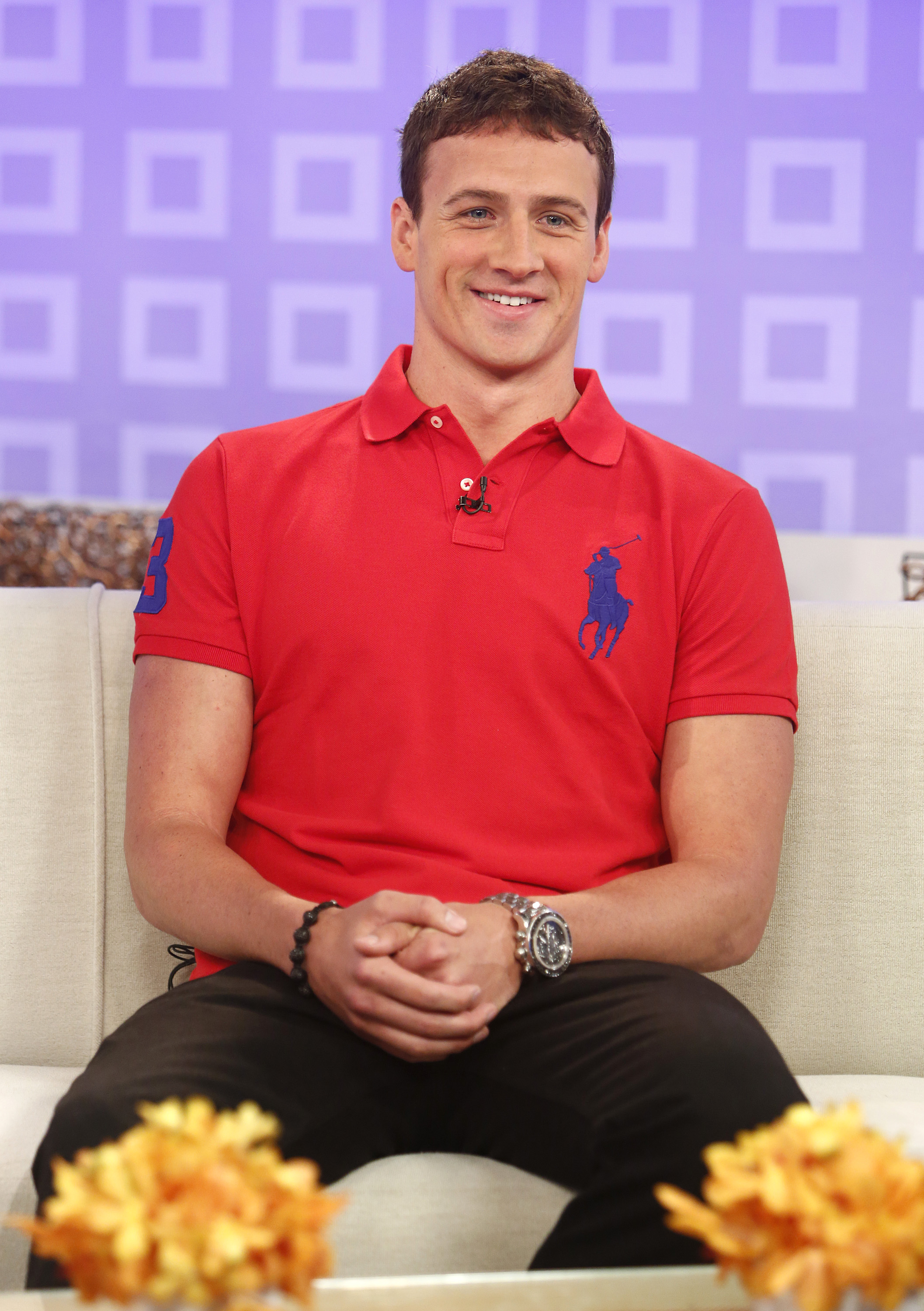 Ryan Lochte appears relaxed while seated on the "Today" show set, wearing a bright red polo shirt paired with dark pants and a silver watch. The Olympic swimmer smiles warmly during his television appearance in front of the studio’s colorful backdrop.