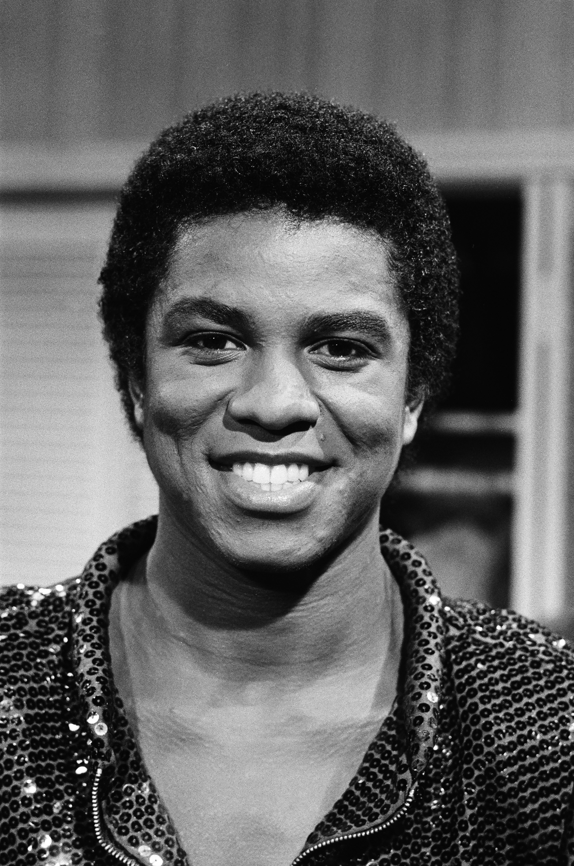 Jermaine Jackson beams in a close-up television appearance, his textured curls and softly shimmering top capturing the polished, performance-ready style of the era. His bright smile and relaxed expression give the moment an easy charm, balancing stage presence with approachability.