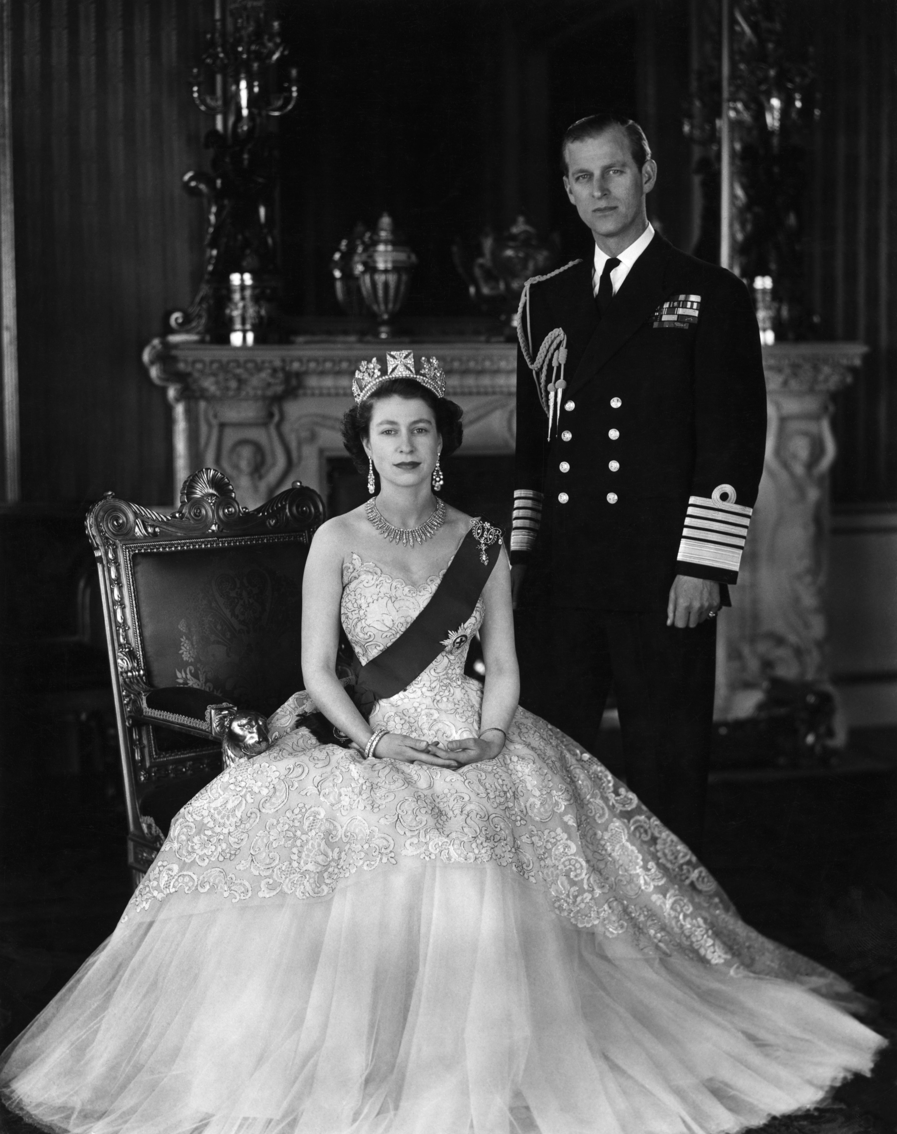 Queen Elizabeth II and Prince Phillip. She is seated and wearing a crown, he is standing in uniform. | Source: Getty Images