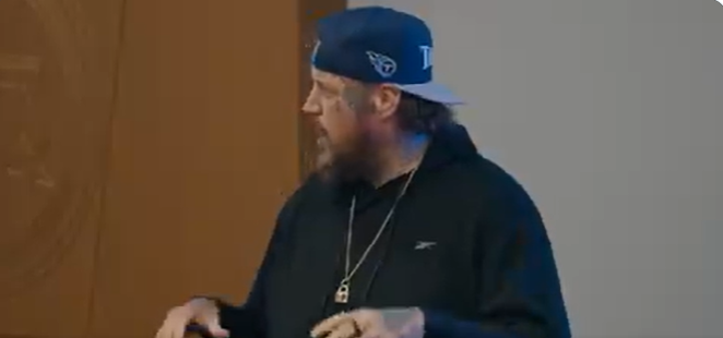 Jason "Jelly Roll" DeFord is captured mid-speech wearing a navy blue Tennessee Titans cap backward, paired with a black Reebok hoodie. A silver pendant necklace rests over his chest as he gestures with one hand, his expression focused and engaged. The casual sportswear look complements his laid-back yet commanding presence in the room | Source: X/TennesseeTitans