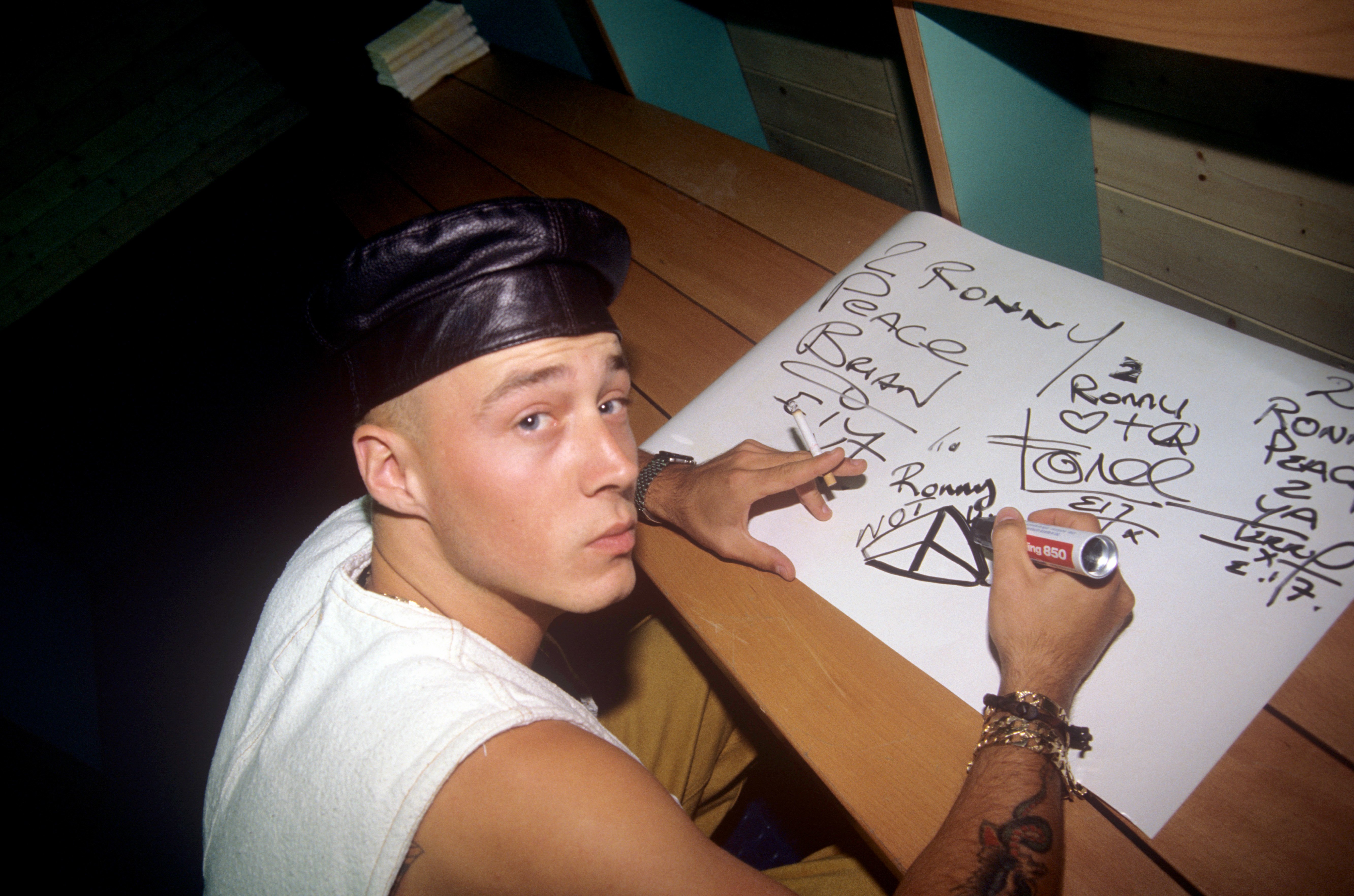 In this candid snapshot taken on 26 June 1993 in Schwenningen, Germany, John Hendy is seen seated at a table, marker in hand, sketching out bold, graffiti-style designs on paper. Wearing a leather cap and layered accessories, the image offers a glimpse into his creative side, blending the band's streetwise aesthetic with a quieter, behind-the-scenes moment from their early rise.