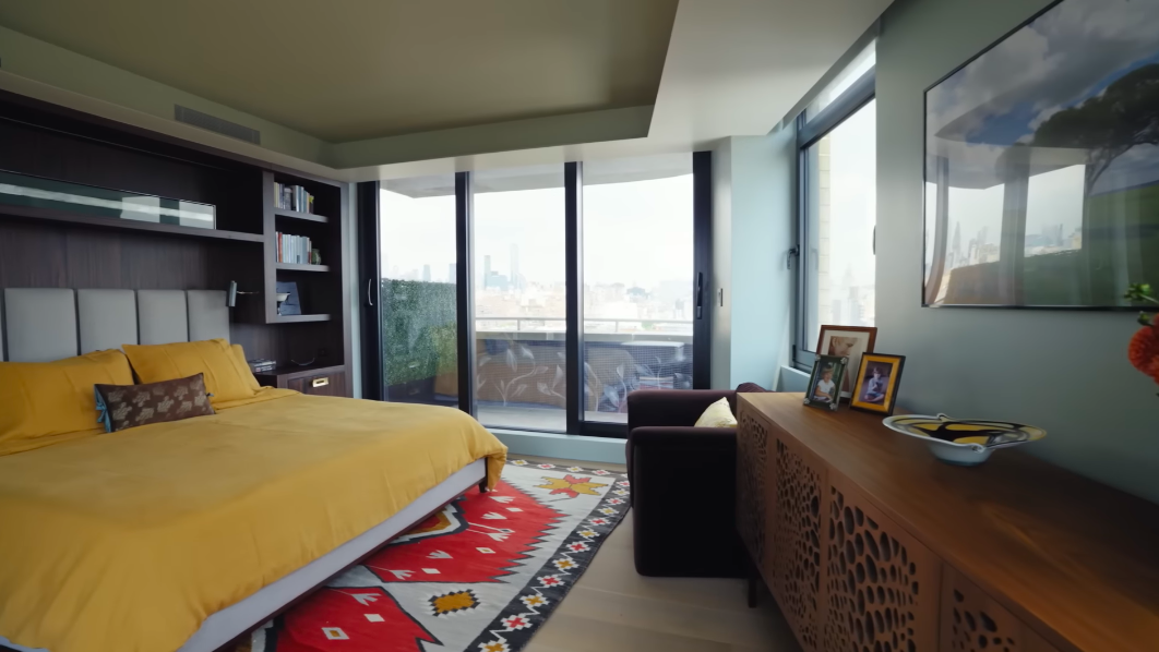 The primary bedroom wraps sleeping space, art and the skyline into one surprisingly warm room. Mustard bedding and a patterned rug stop those expansive windows from making the space feel cold. | Source: YouTube/Architectural Digest