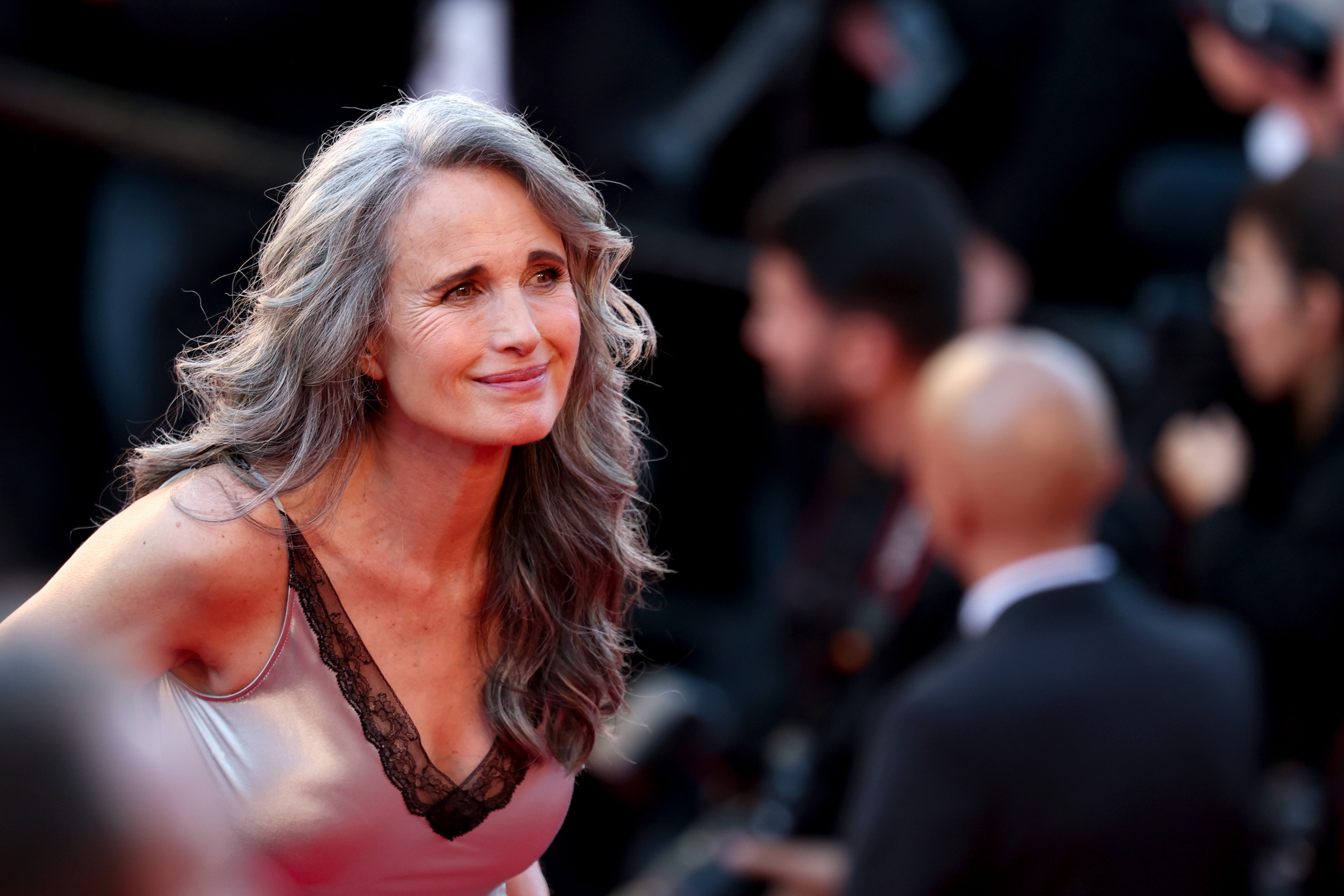 Andie MacDowell leans slightly forward with a warm smile, acknowledging someone off-camera as she attends the "Dossier 137" red carpet during Cannes. Her gray hair and silver gown glow under the spotlight.