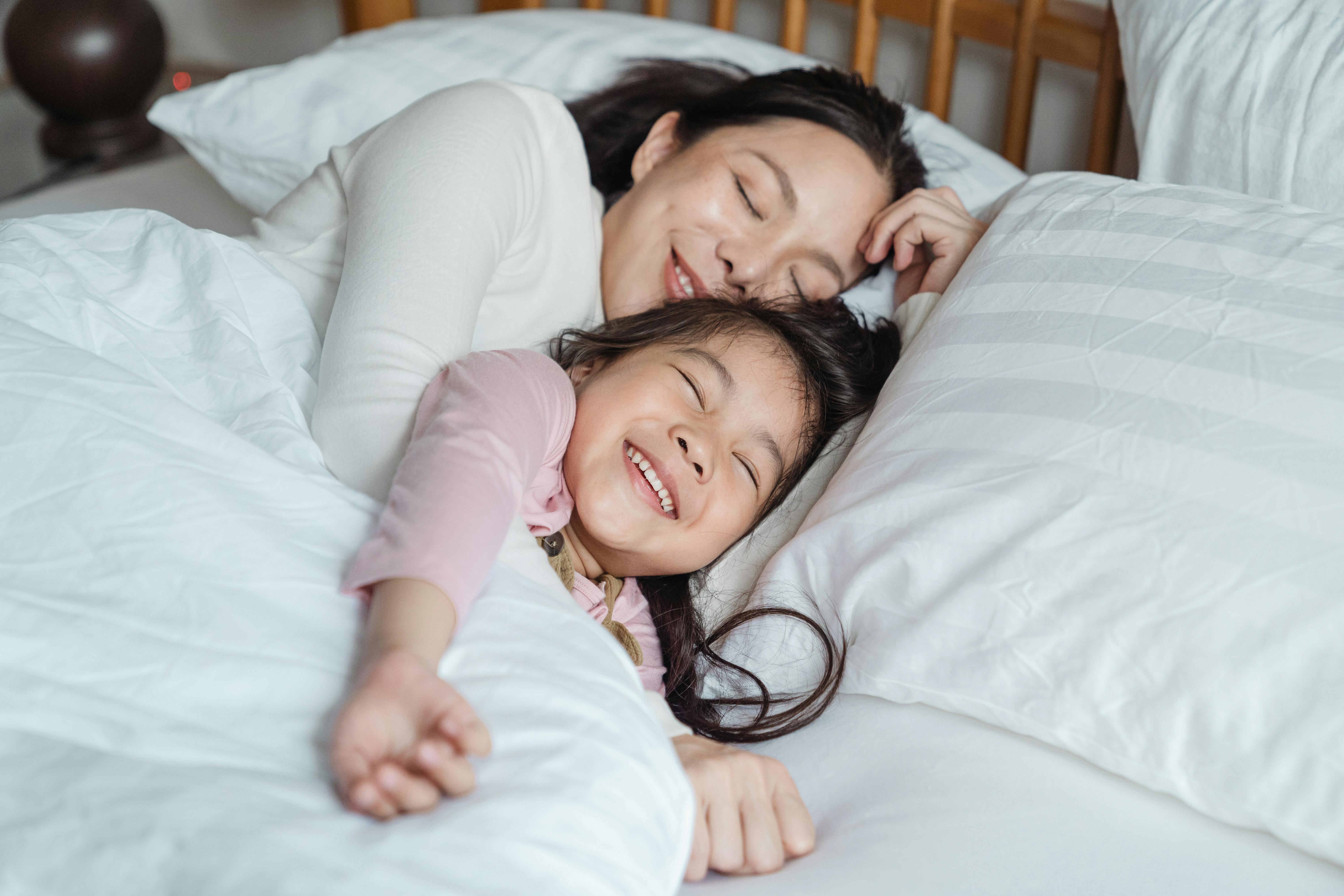 A woman smiling while comforting a little girl while they lay in a bed together | Source: Pexels