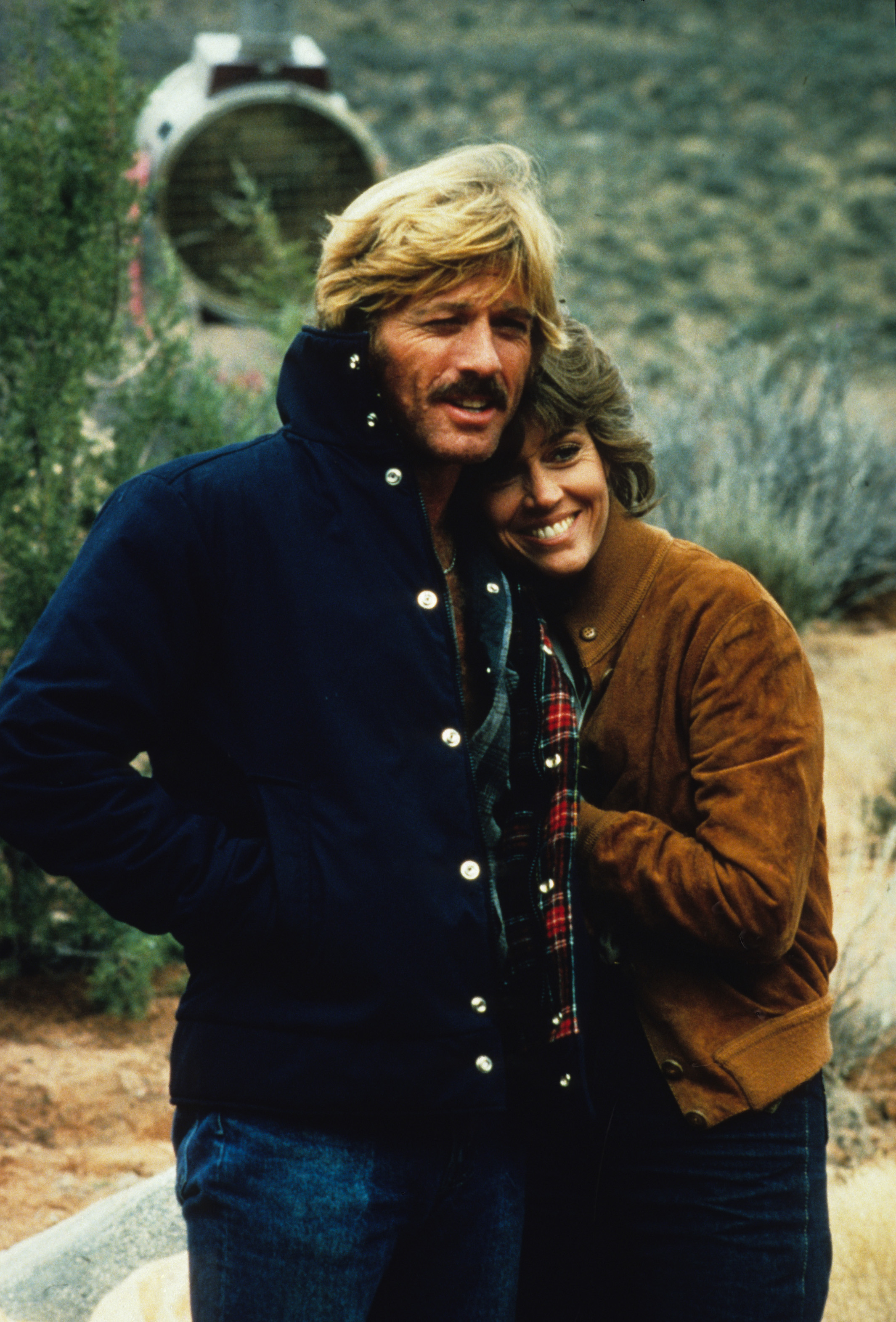 Still on the set of the film "The Electric Horseman," directed by Sydney Pollack, Robert Redford and Jane Fonda appear at ease, with Fonda resting her head on Redford's shoulder as they smile happily.
