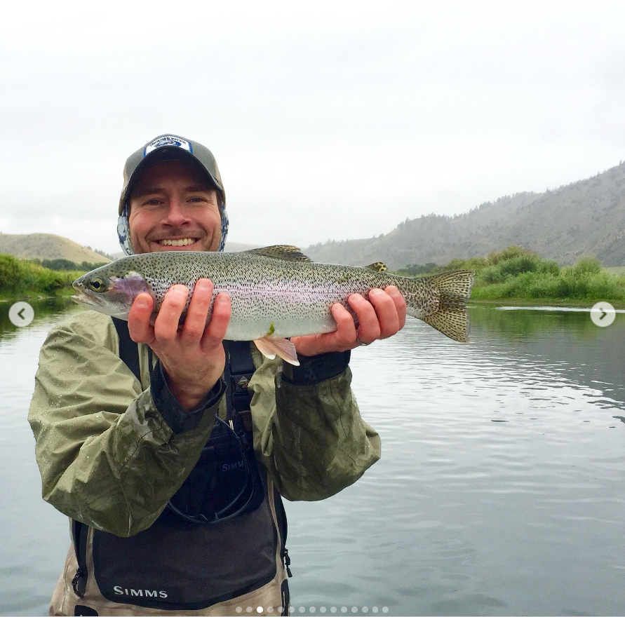 Matthew Mitchell smiles while holding up a freshly caught fish against a calm river and misty mountain backdrop. | Source: Instagram/thy_travelers