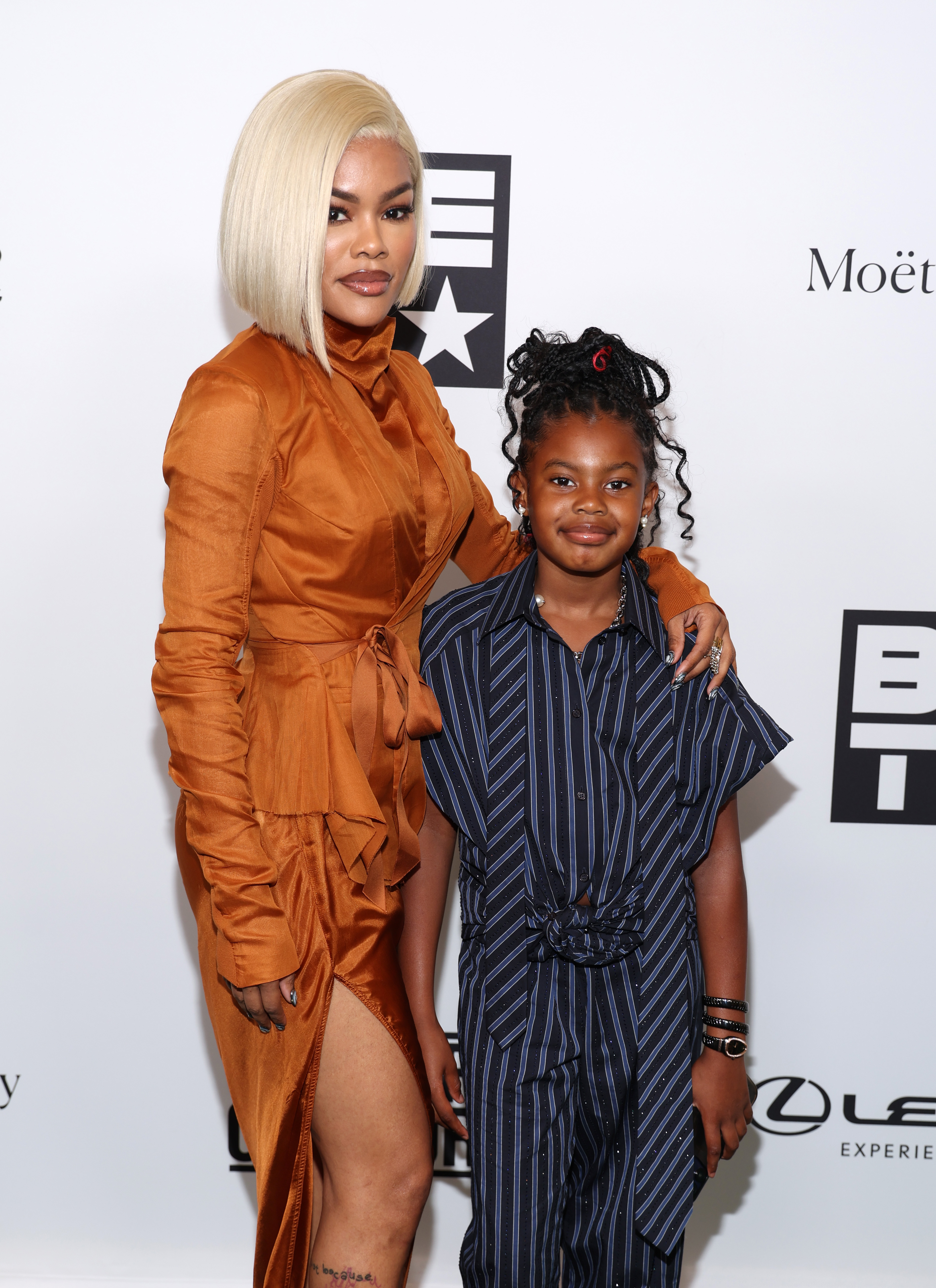 Teyana Taylor poses with Iman Tayla "Junie" Shumpert Jr. at the Culture Creators brunch in Los Angeles. Teyana wears an orange dress while Junie smiles beside her in a striped outfit.
