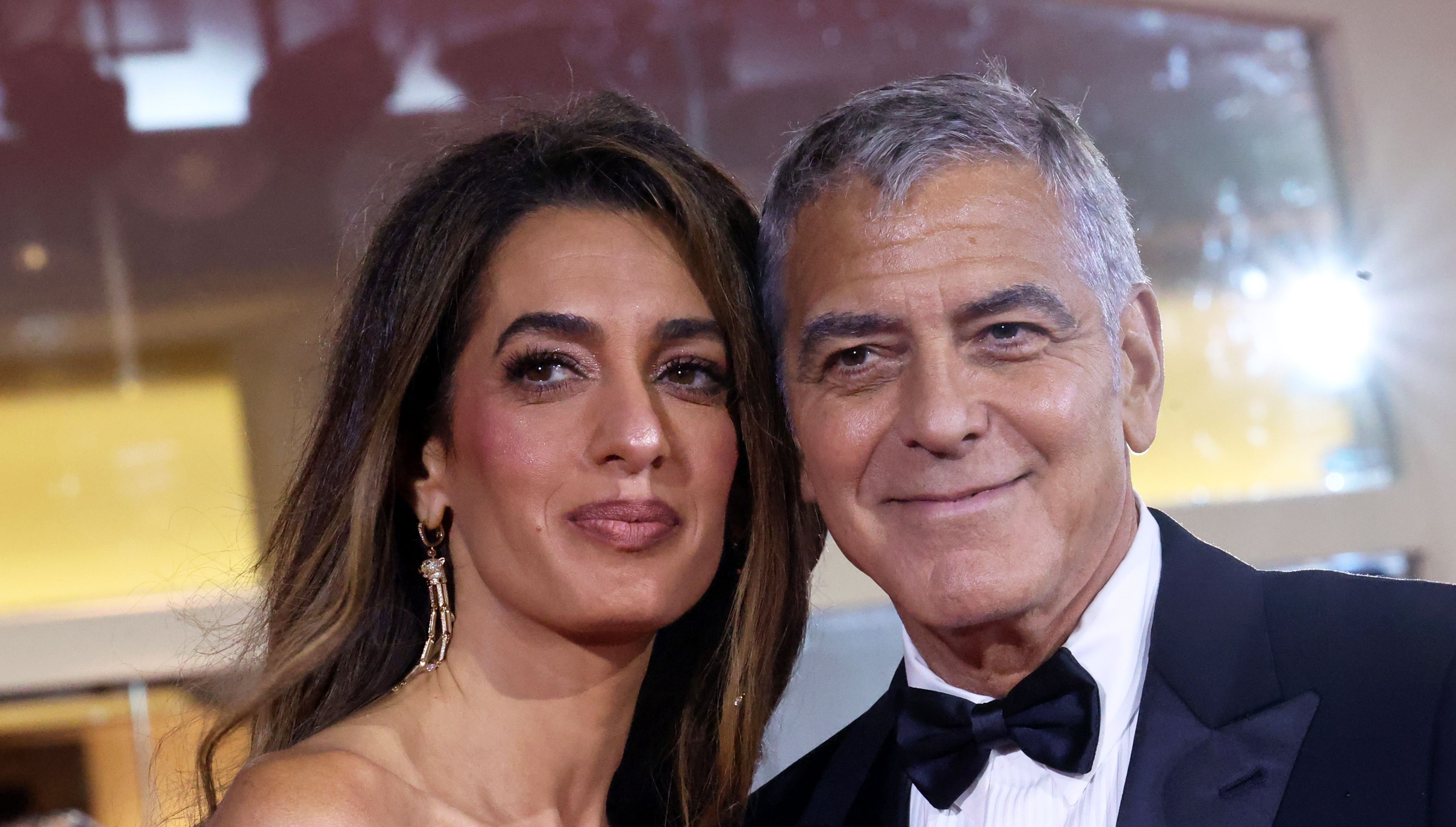 Amal and George Clooney pose closely for the cameras, both smiling softly in this intimate close-up. Amal's glowing makeup and statement earrings complement her effortless waves, while George's bow tie and tailored tux complete the polished look.