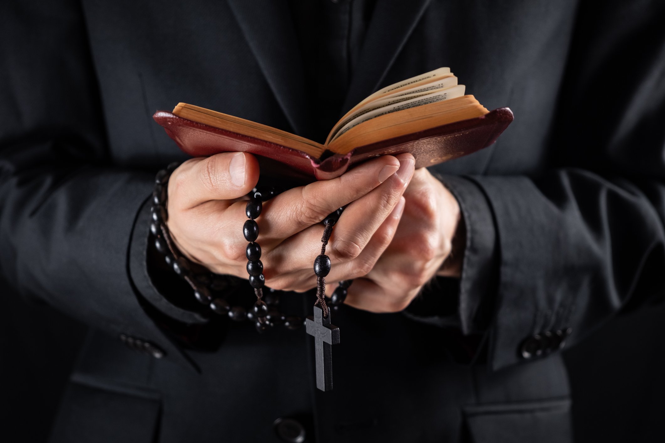 Hands of a christian priest dressed in black holding a crucifix and reading New Testament book | Photo: Getty Images