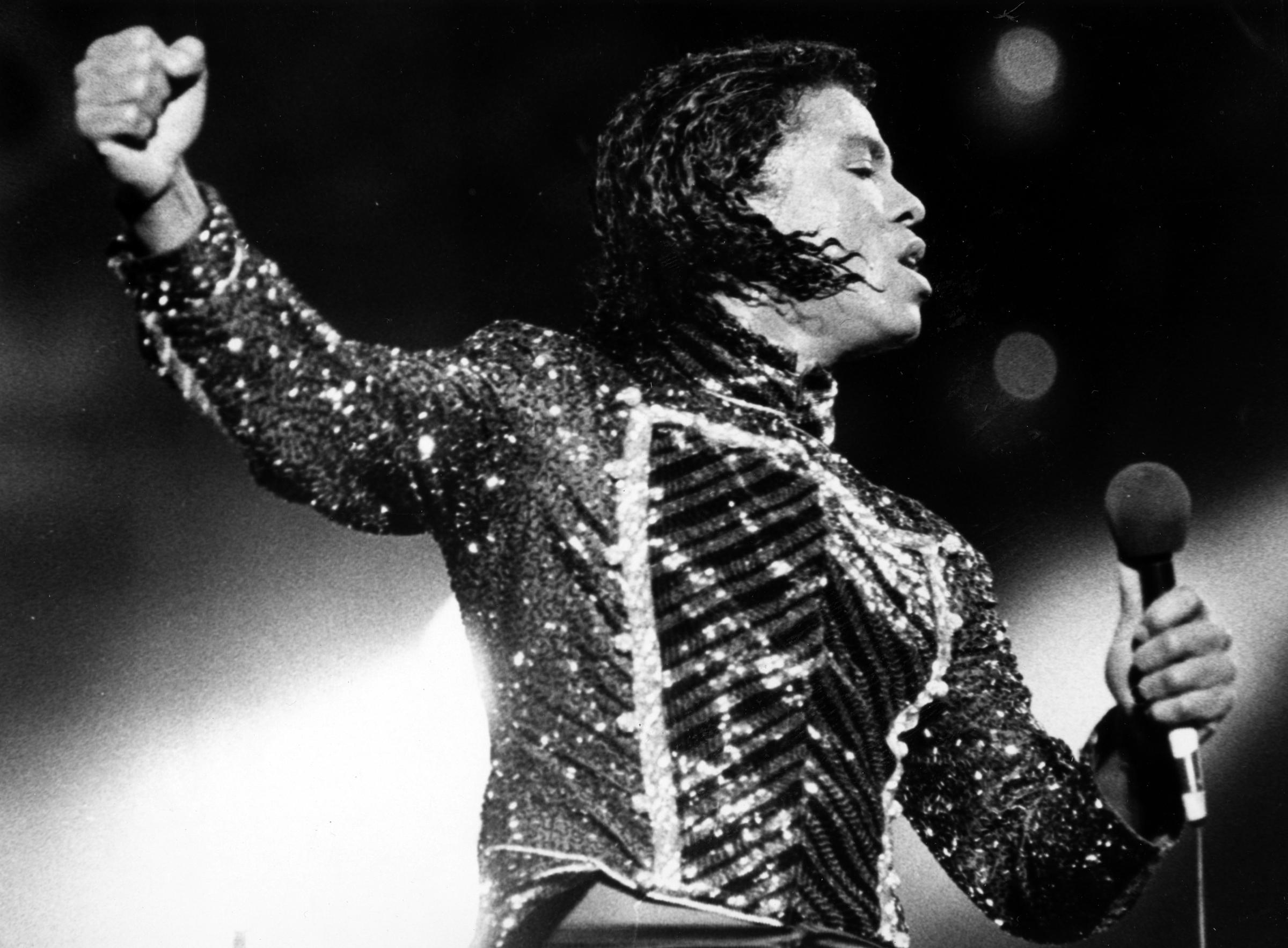 Jermaine Jackson takes the stage in a glittering, form-fitting jumpsuit, its bold, shimmering pattern catching the light as he performs. With microphone in hand and arm raised, he delivers a high-energy moment that reflects the showmanship of the era.