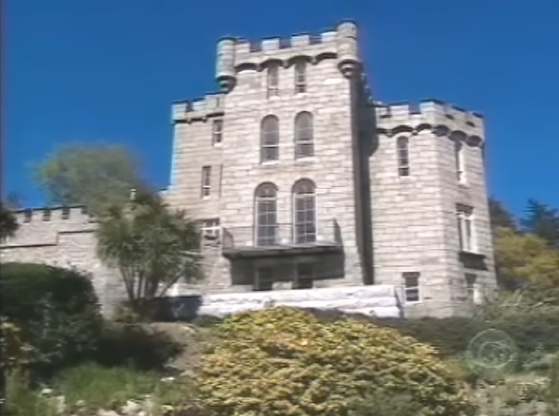 A closer exterior view highlights the symmetry of the 19th-century structure, from its tall, arched windows to the sturdy corner towers anchoring either side. Bathed in bright daylight, the castle's stonework and elevated position reflect the commanding yet peaceful setting Enya chose to call home. | Source: YouTube/AndreW