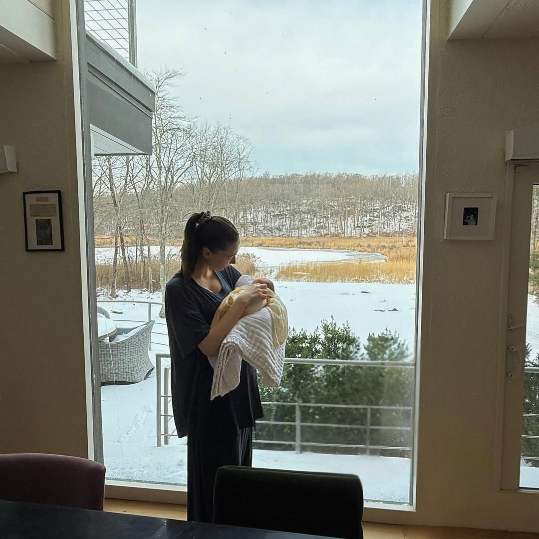 Elsie Hewitt stands by a snowy window holding baby Scottie Rose in a quiet moment at home, as posted on December 18, 2025 | Source: Instagram/elsie