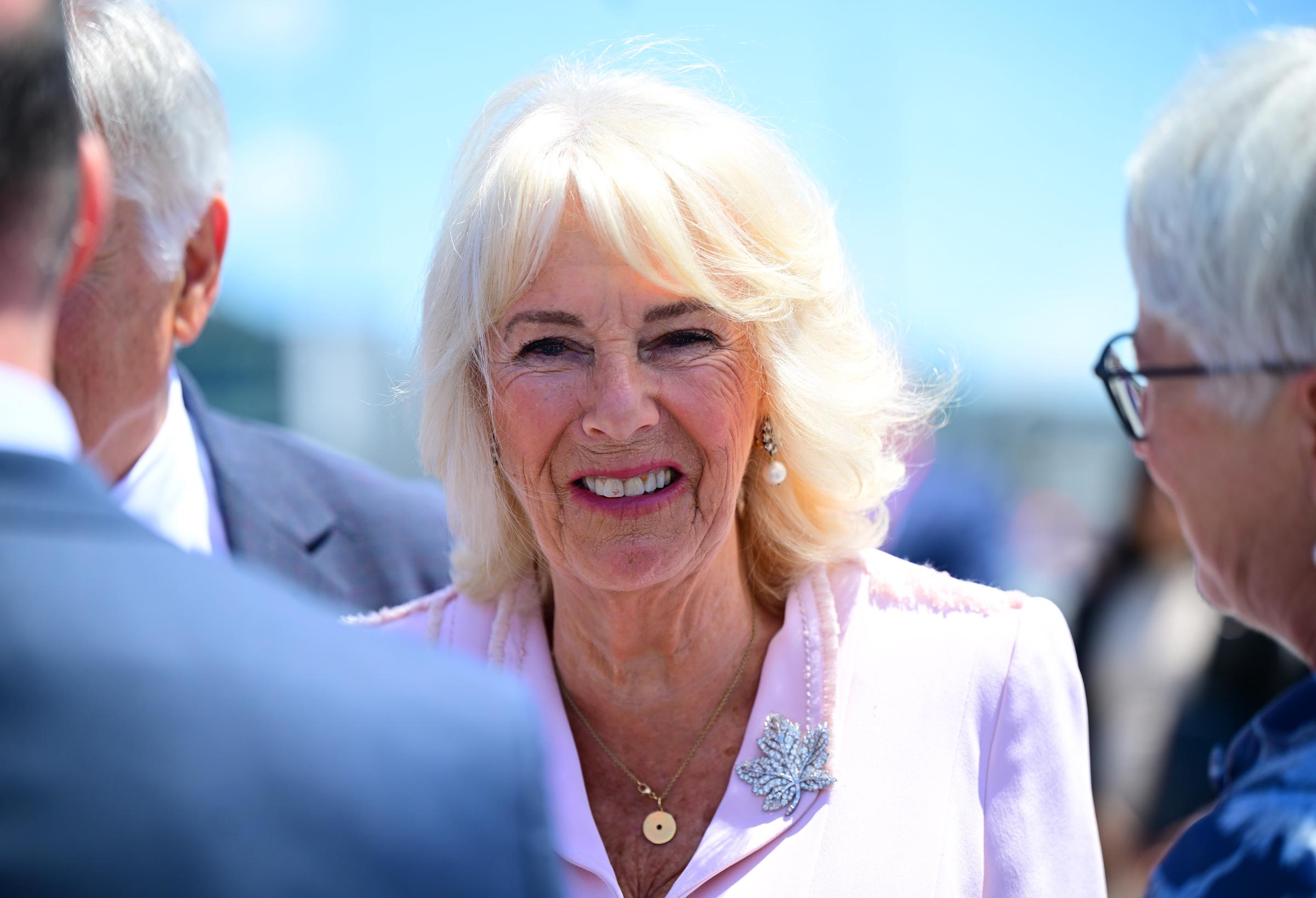 The camera focuses on Queen Camilla as she smiles while greeting gathered officials. She wears a diamond maple leaf brooch, pearl drop earrings, and a gold necklace.