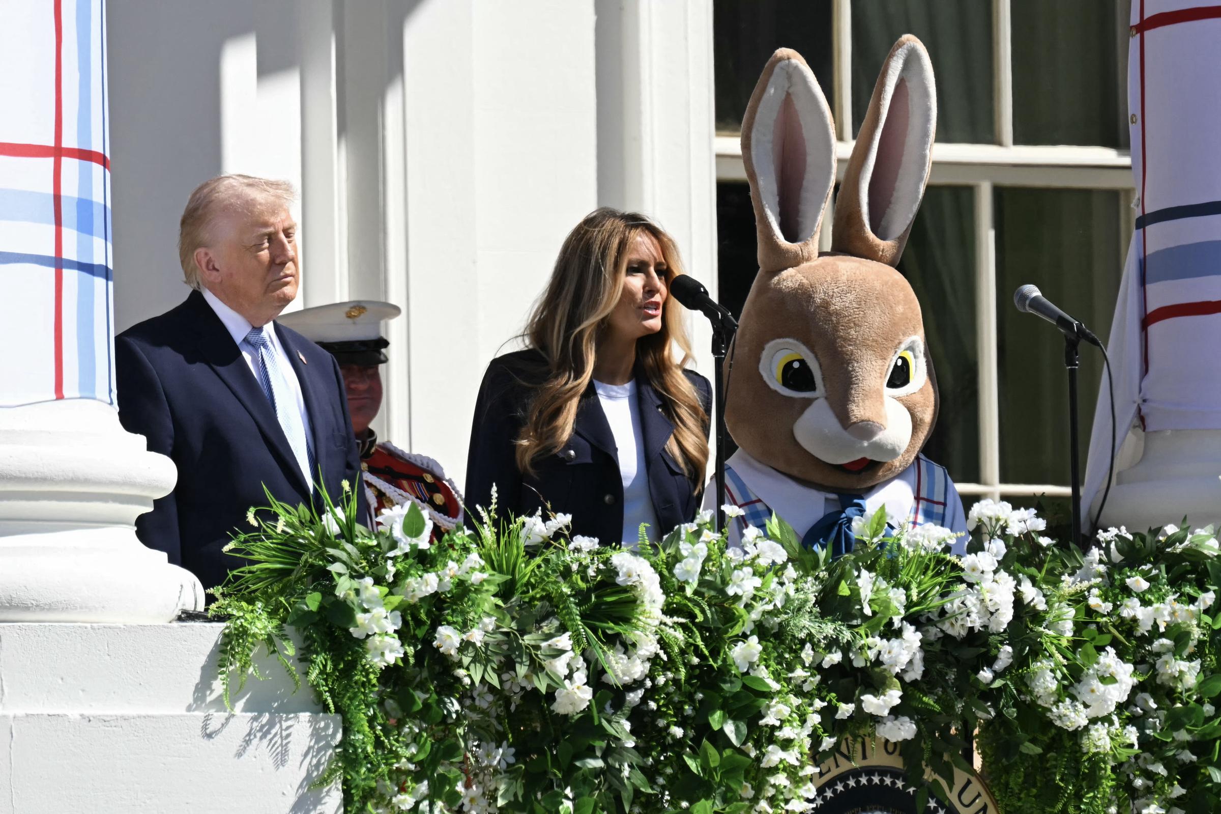 Melania Trump speaks from the White House balcony as Donald Trump and the Easter Bunny stand beside her, surrounded by cascading greenery and white blooms.