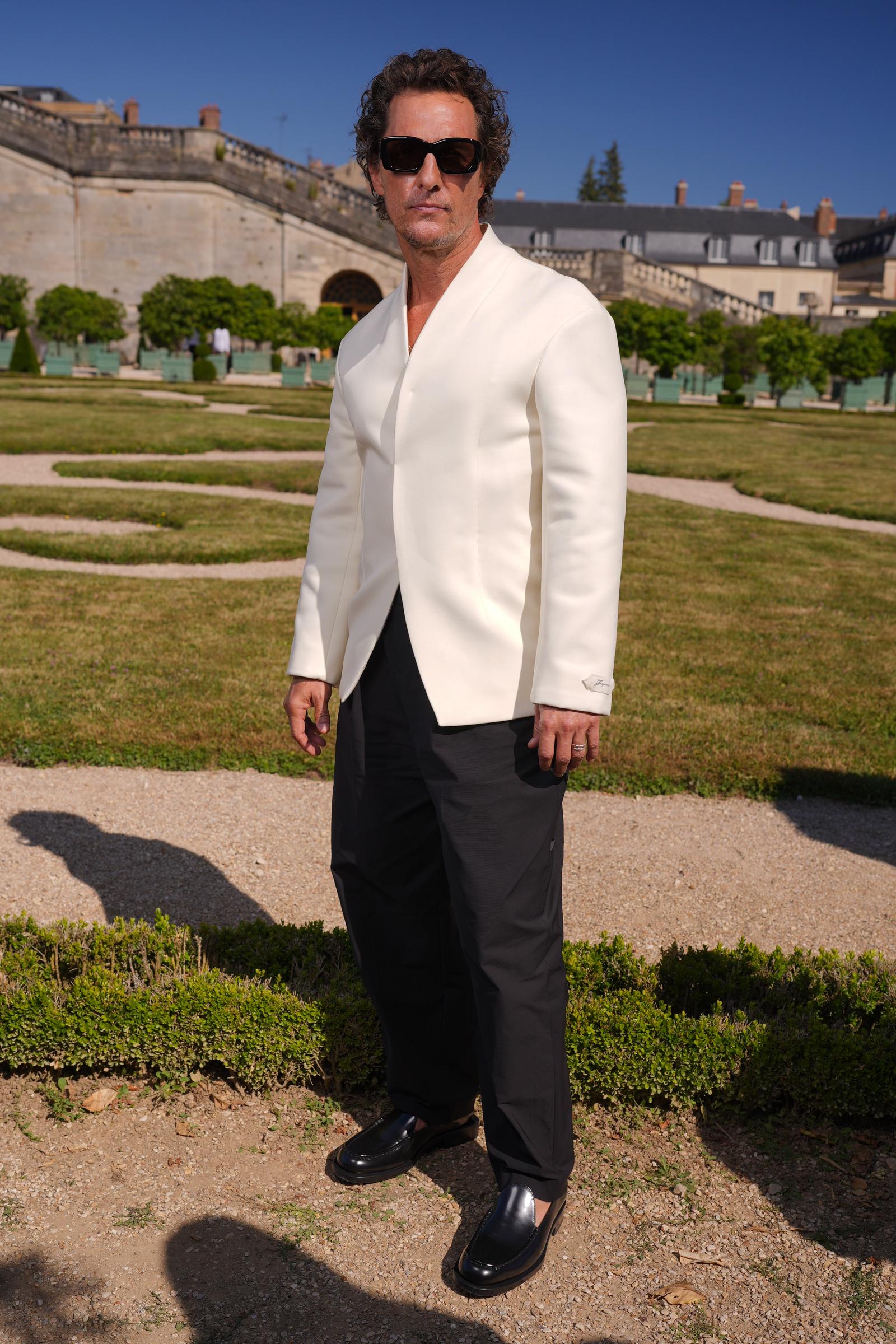McConaughey poses outside the Château de Versailles, exuding effortless style. He wears a structured cream jacket with a minimalist, collarless cut, paired with black pleated trousers and black leather loafers. His look is completed with rectangular black sunglasses and layered rings, adding a touch of modern edge to the classic ensemble.
