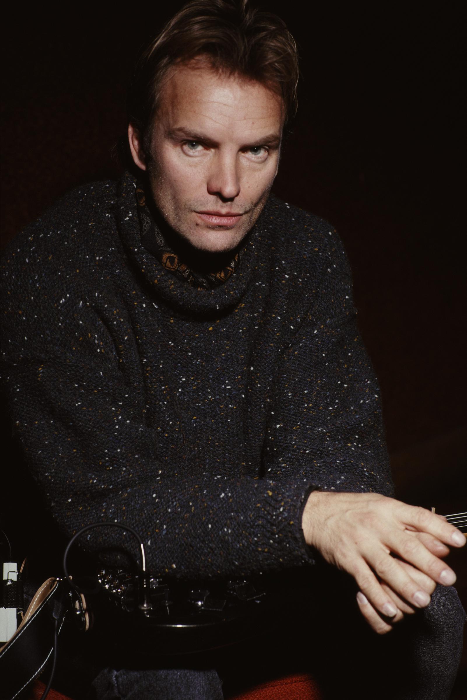 A moody studio portrait of Sting in New York, January 1991 — leaning forward with arms resting on a guitar, wearing a speckled dark turtleneck, his blue eyes fixed intently on the camera.