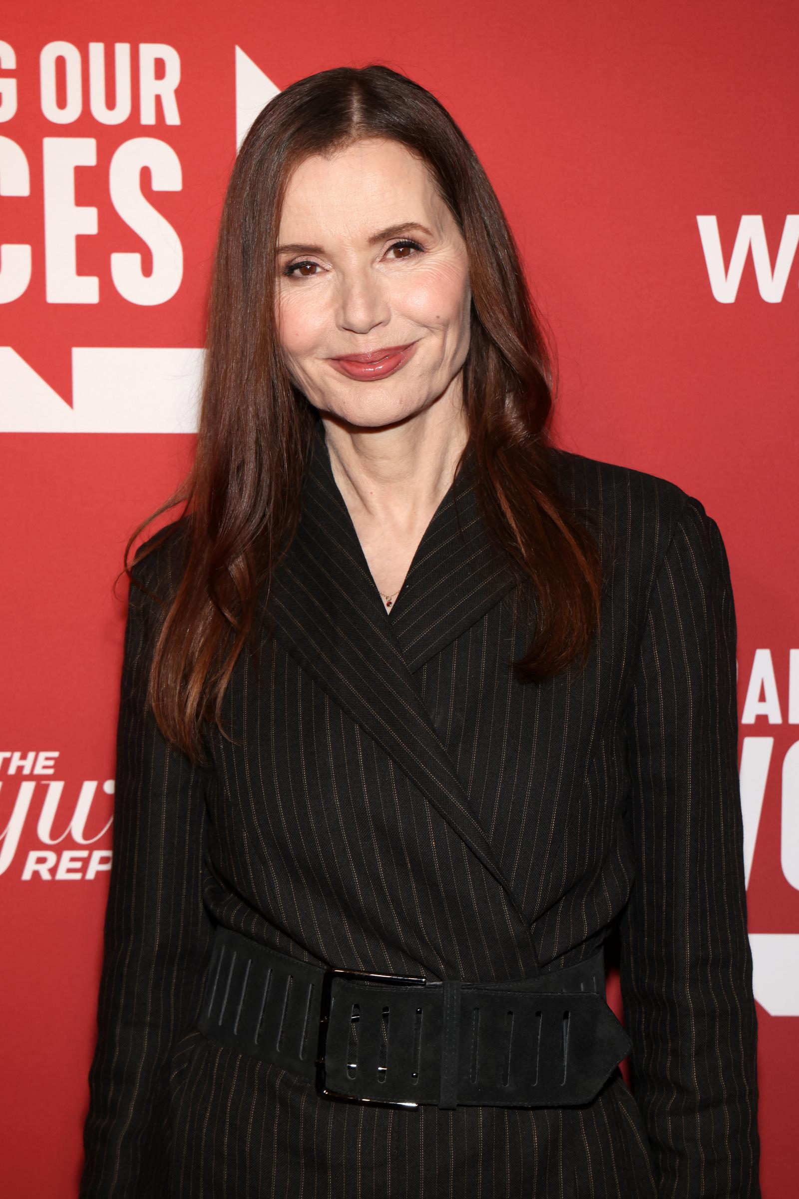 Geena Davis arrived in a tailored pinstriped blazer dress cinched with a dramatic wide belt, blending power dressing with understated glamour. Her sleek straight hair and subtle smile gave the look a confident, polished finish against the striking red backdrop.