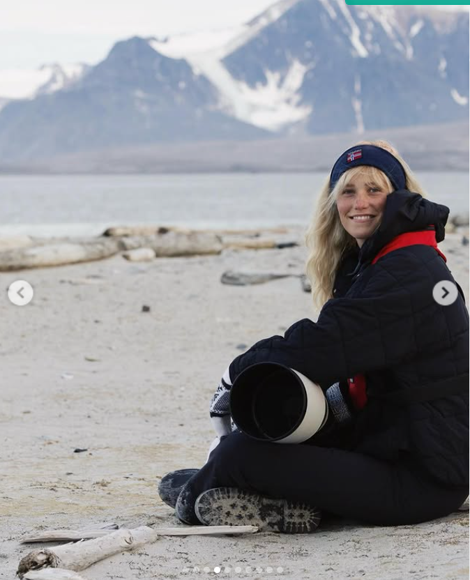 Asleigh Scully sits on a windswept shore, camera in hand, with snow-capped mountains behind her. | Source: Instagram/ascullyphoto