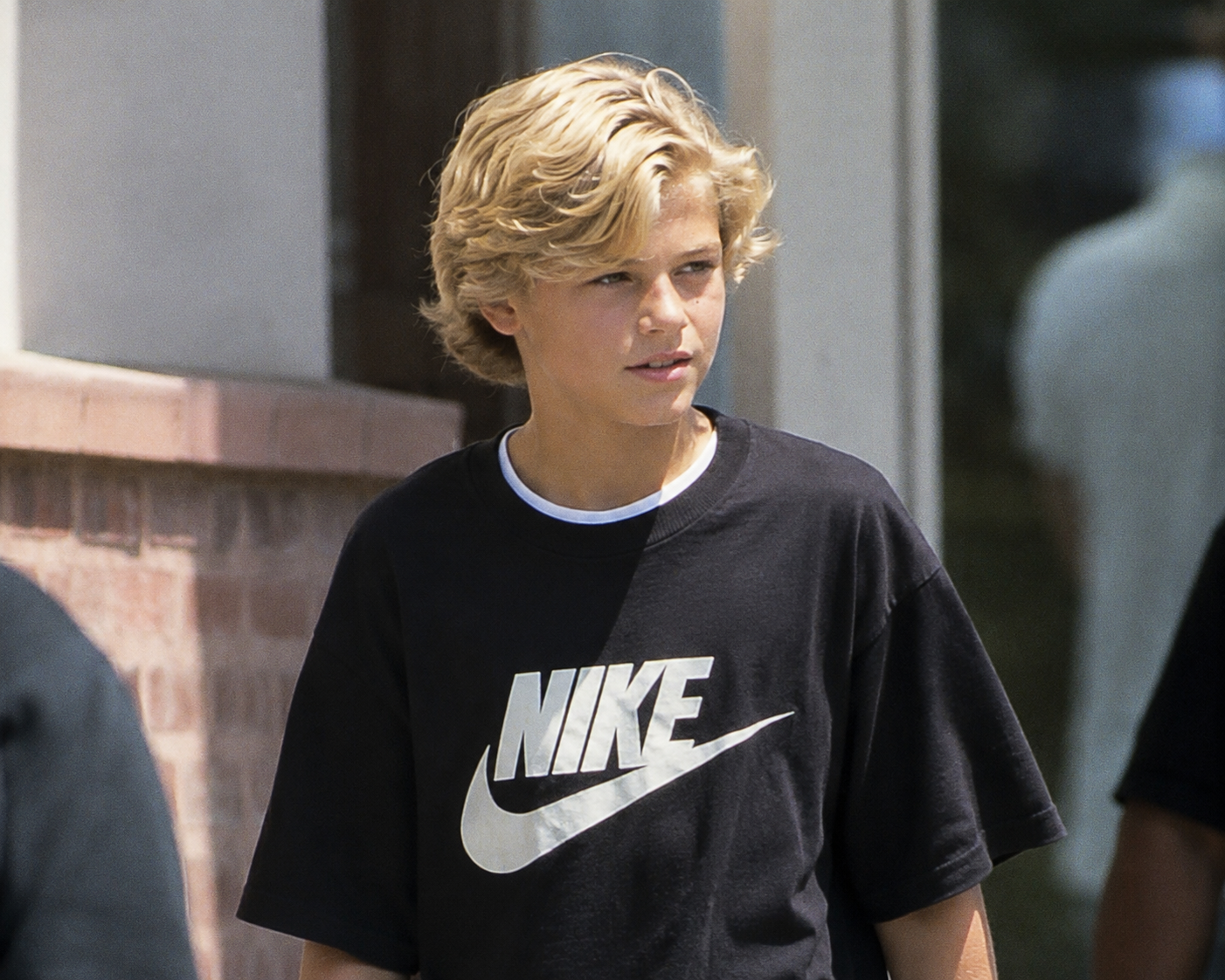 In this AI-imagined shot, the older son walks outside wearing a black Nike graphic tee, capturing a candid early 2000s street-style moment. | Source: ChatGPT