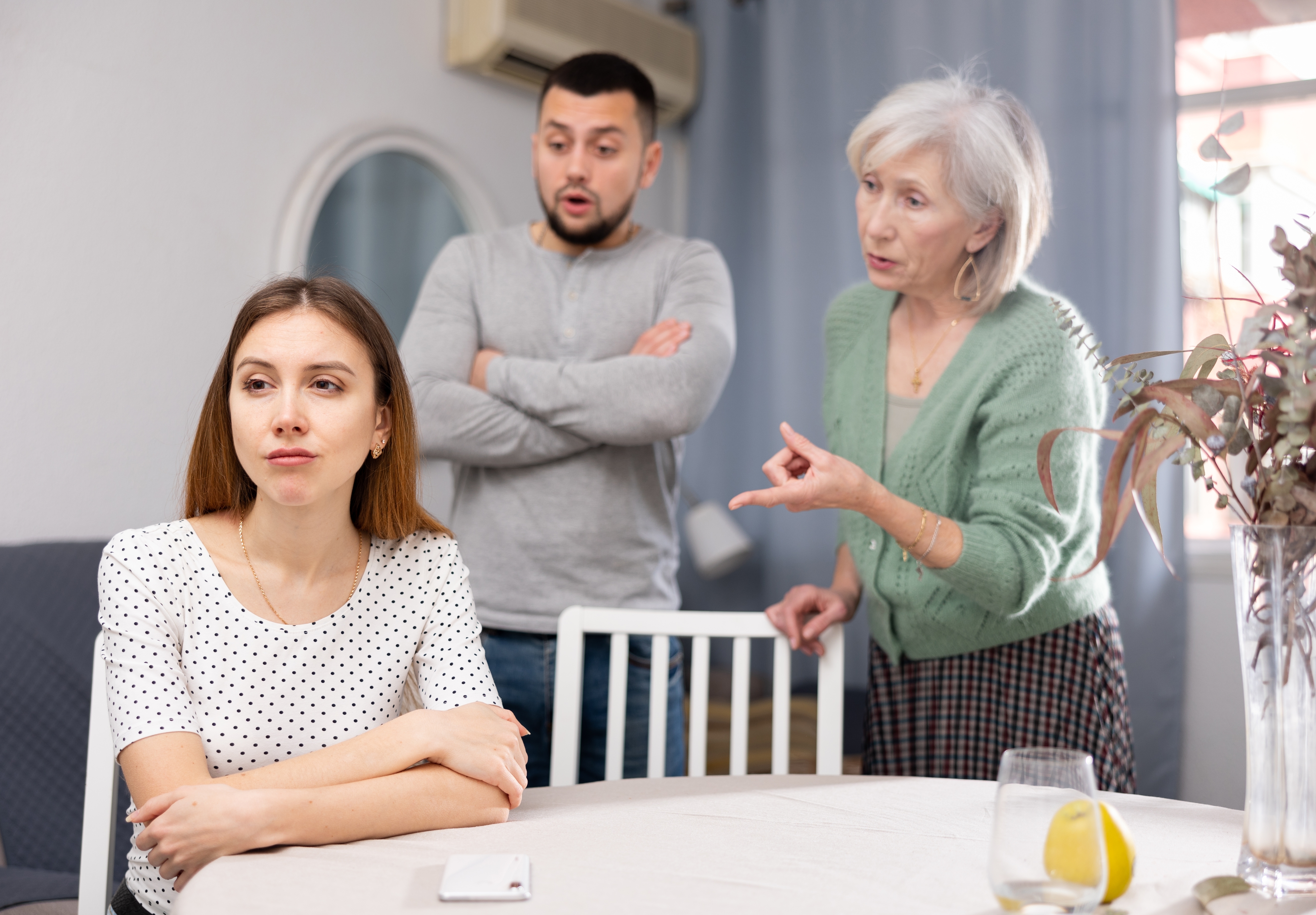 A woman sitting down looking upset while her mother-in-law stands behind her scolding while her son stands beside her | Source: Shutterstock