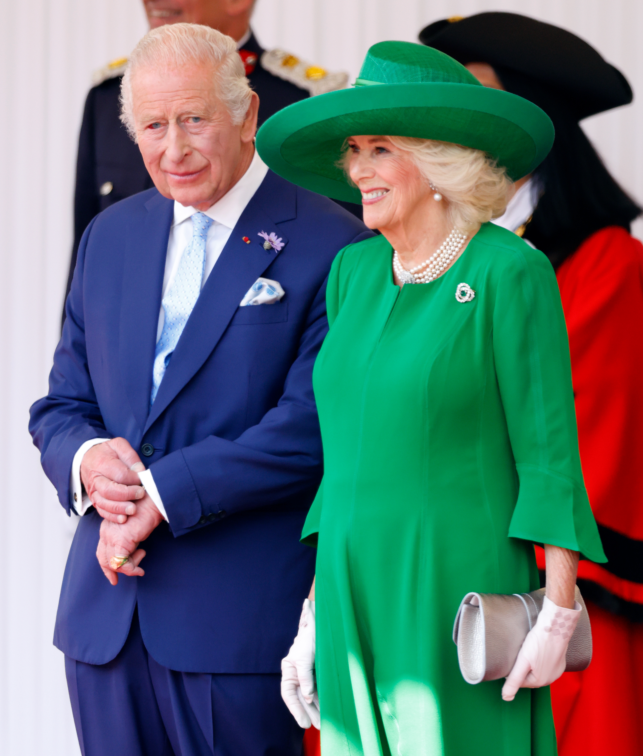 King Charles III and Queen Camilla attend the formal welcome for French President Emmanuel Macron and Brigitte Macron's 2025 state visit to the United Kingdom. The king, dressed in royal blue with a visible bloodshot eye, stands beside his wife, who stuns in vibrant green and pearls at the Royal Dais in Windsor.