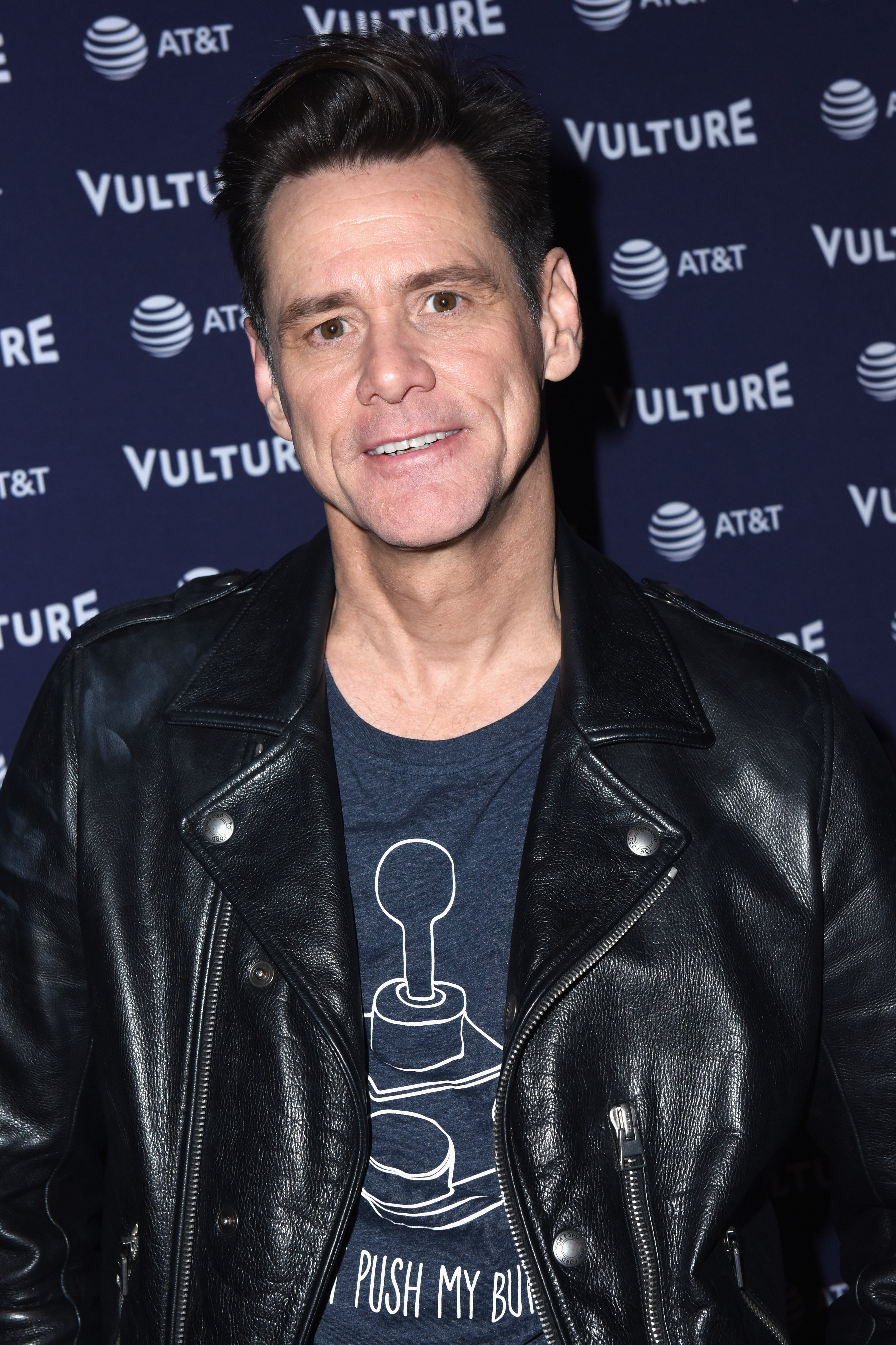 Jim Carrey attends "Jim Carrey In Conversation with Jerry Saltz" during the Vulture Festival presented by AT&T. He wears a black leather jacket over a graphic T-shirt, smiling warmly at the camera. The event backdrop featuring the Vulture and AT&T logos adds a professional yet relaxed atmosphere.