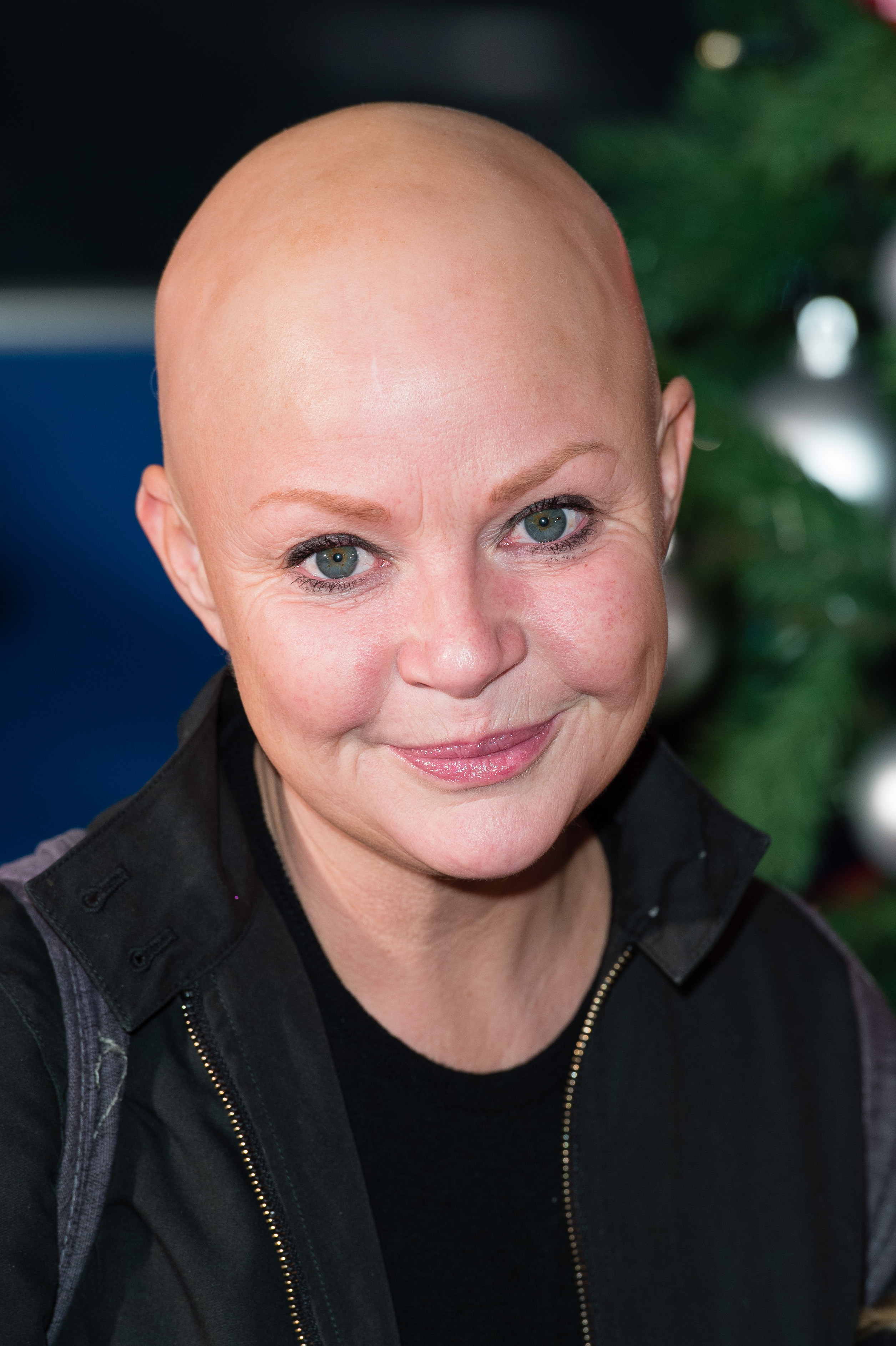 A close portrait of Porter, fully bald and without eyebrows, her pale green eyes the focal point of the image against a softly blurred backdrop of Christmas greenery and silver baubles. She wears a black zip-up jacket and a gentle smile. Pictured at the VIP preview of Winter Wonderland, Hyde Park, London, November 2016.