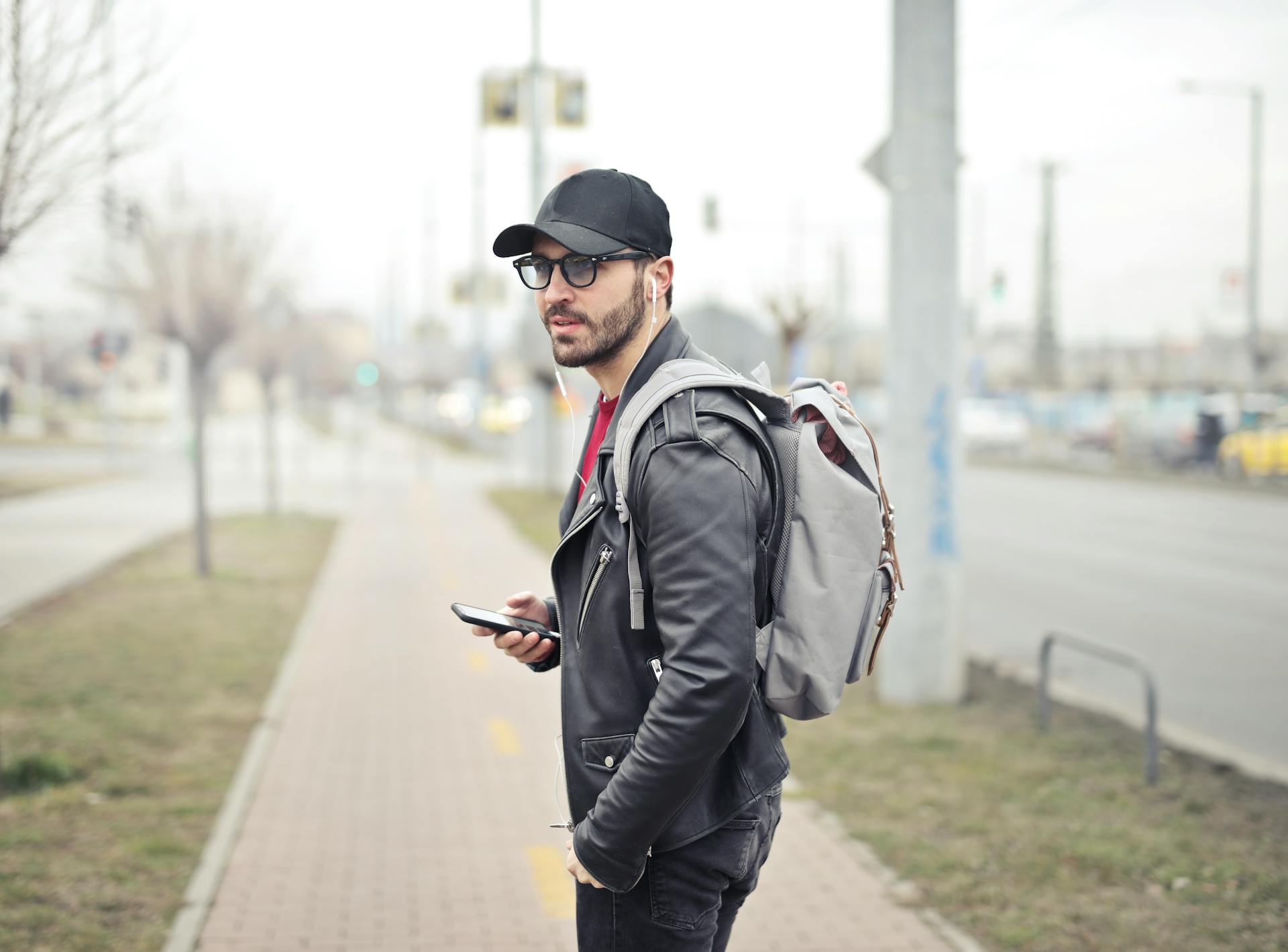 A man in a black jacket and a backpack holding a phone while walking on the sidewalk | Source: Pexels