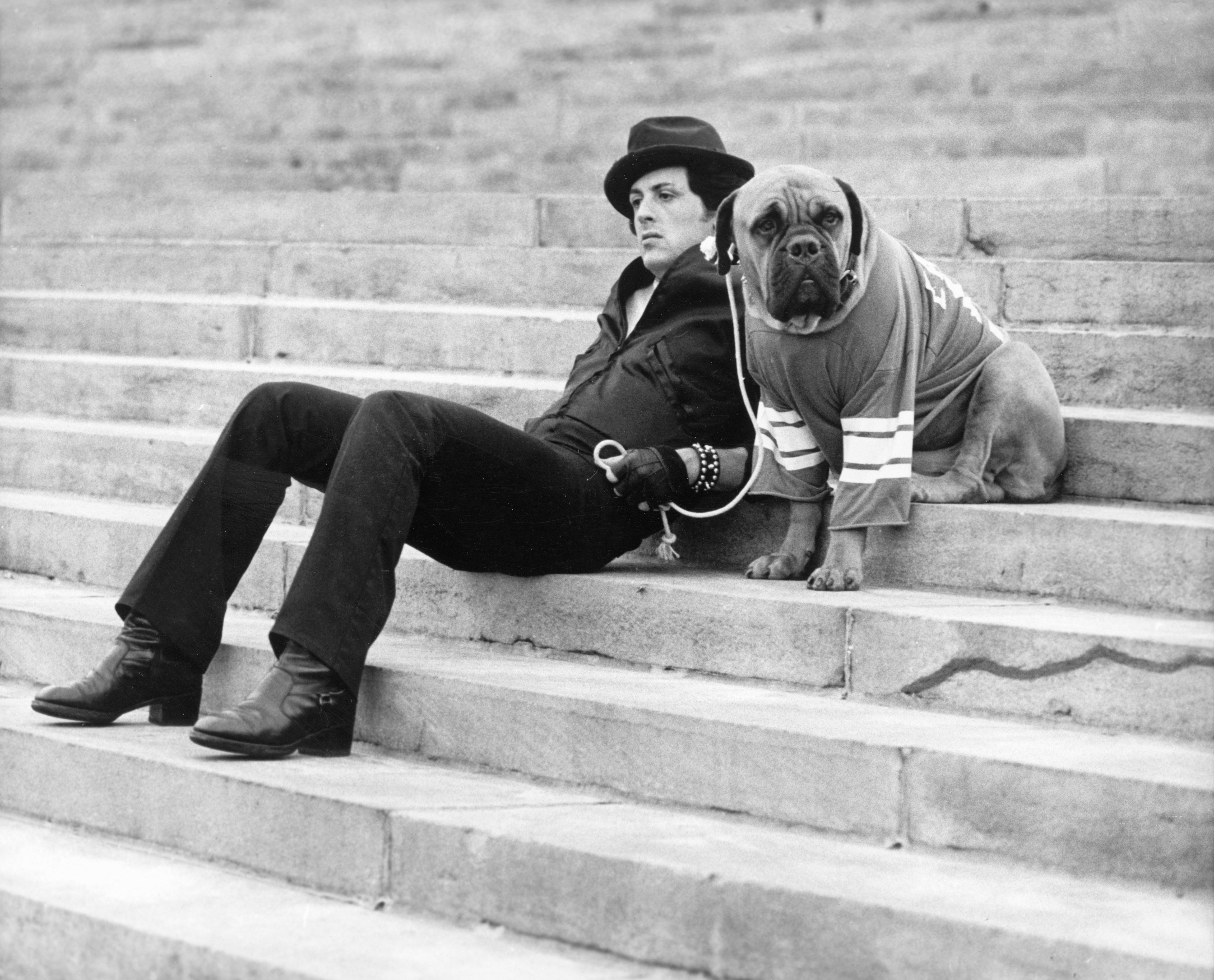 Sylvester Stallone sits on a staircase while holding the leash of a dog in a still from the film, "Rocky" in 1976. | Photo: Getty Images.