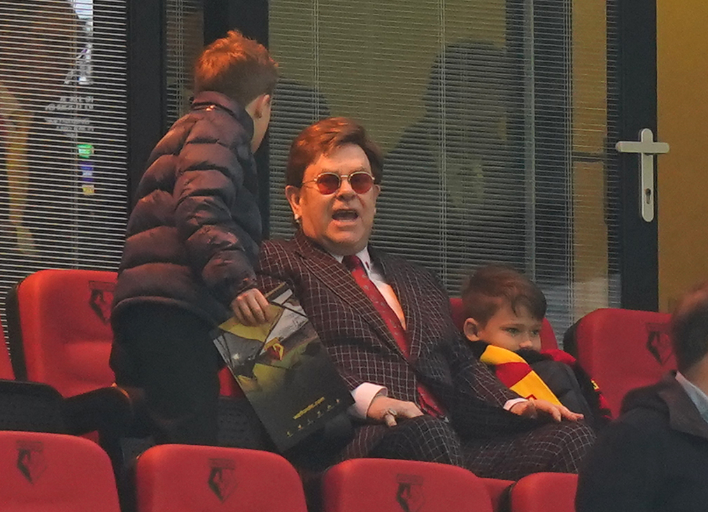 Sir Elton John took his seat in the stands at Vicarage Road on May 8, 2021, flanked by Zachary John Furnish-John and Elijah Joseph Daniel Furnish-John ahead of the Sky Bet Championship match in England. Dressed in a sharp checked suit with a bold red tie and his trademark tinted glasses, the singer looked animated as one son leaned in beside him while the other sat wrapped in a red-and-yellow scarf.