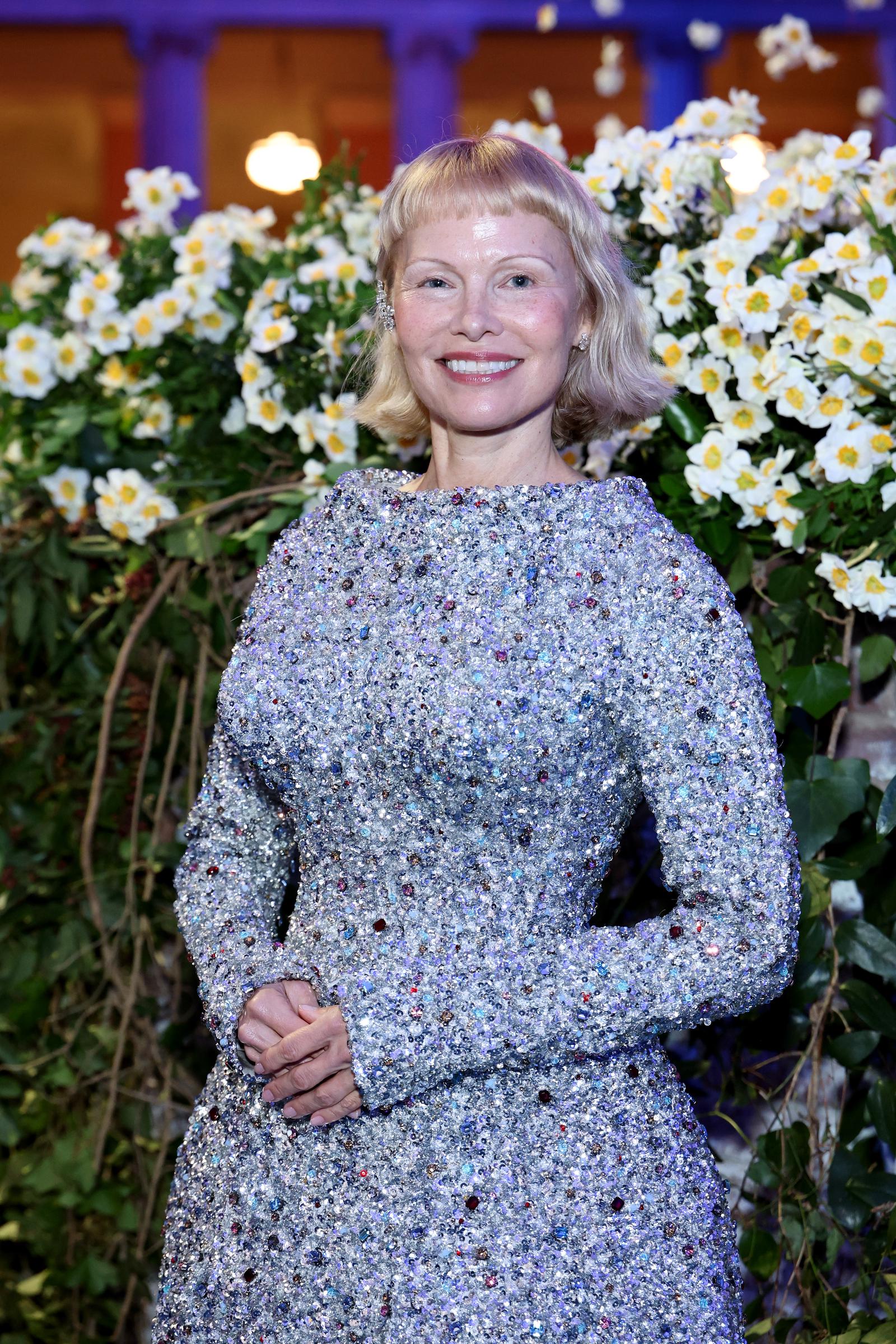 Framed by a wall of white blossoms, Pamela Anderson beams during a quiet moment at the 2025 Met Gala. Her fresh-faced appearance and custom Tory Burch gown echoed the event's tribute to elegant tailoring and craftsmanship.