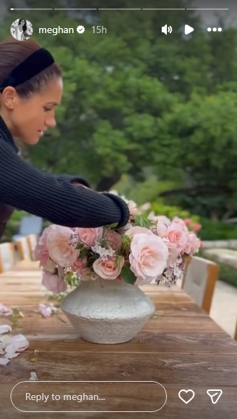 Meghan leans over a large stone-effect vase brimming with soft pink roses, carefully arranging the blooms on a wooden outdoor dining table at the Montecito estate, scattered petals resting on the surface beside her. She is dressed in a dark long-sleeved top and a black headband, with a lush canopy of green trees filling the background. | Source: Instagram/meghan