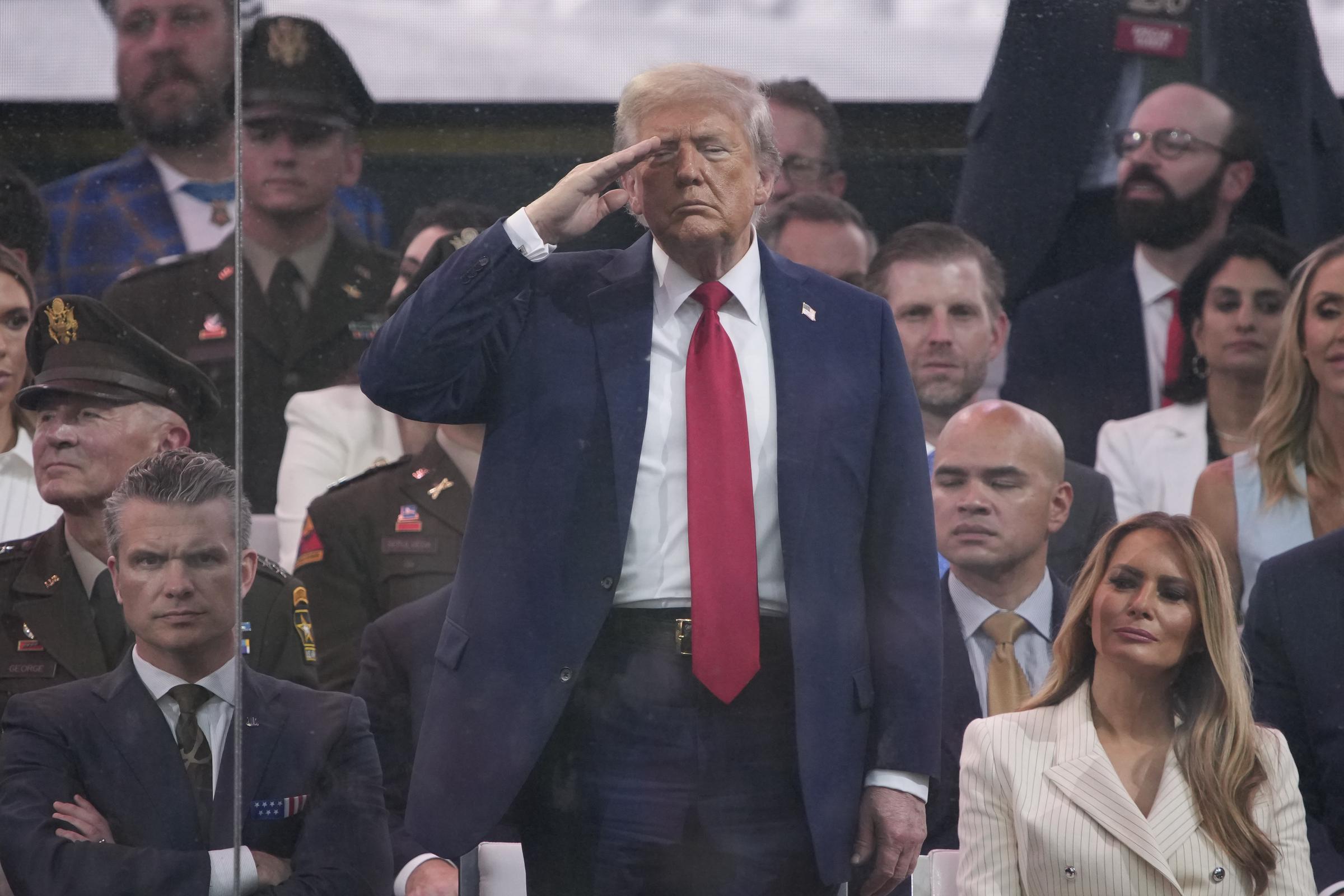 Donald Trump stands and salutes while members of the US Army march by during the parade. Melania sits beside him in the front row, and the moment captures a solemn tribute as military officials look on.