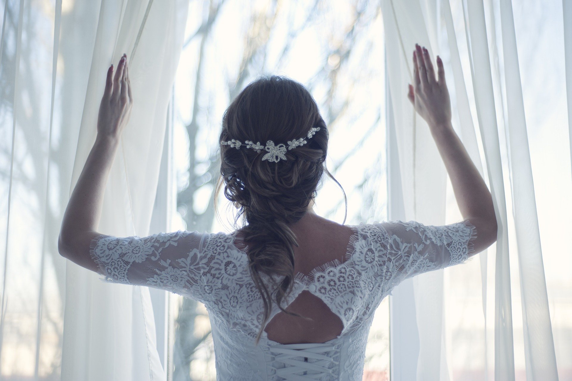 Eine Braut in einem Spitzenhochzeitskleid, das an einem großen Fenster steht. | Quelle: Pixabay