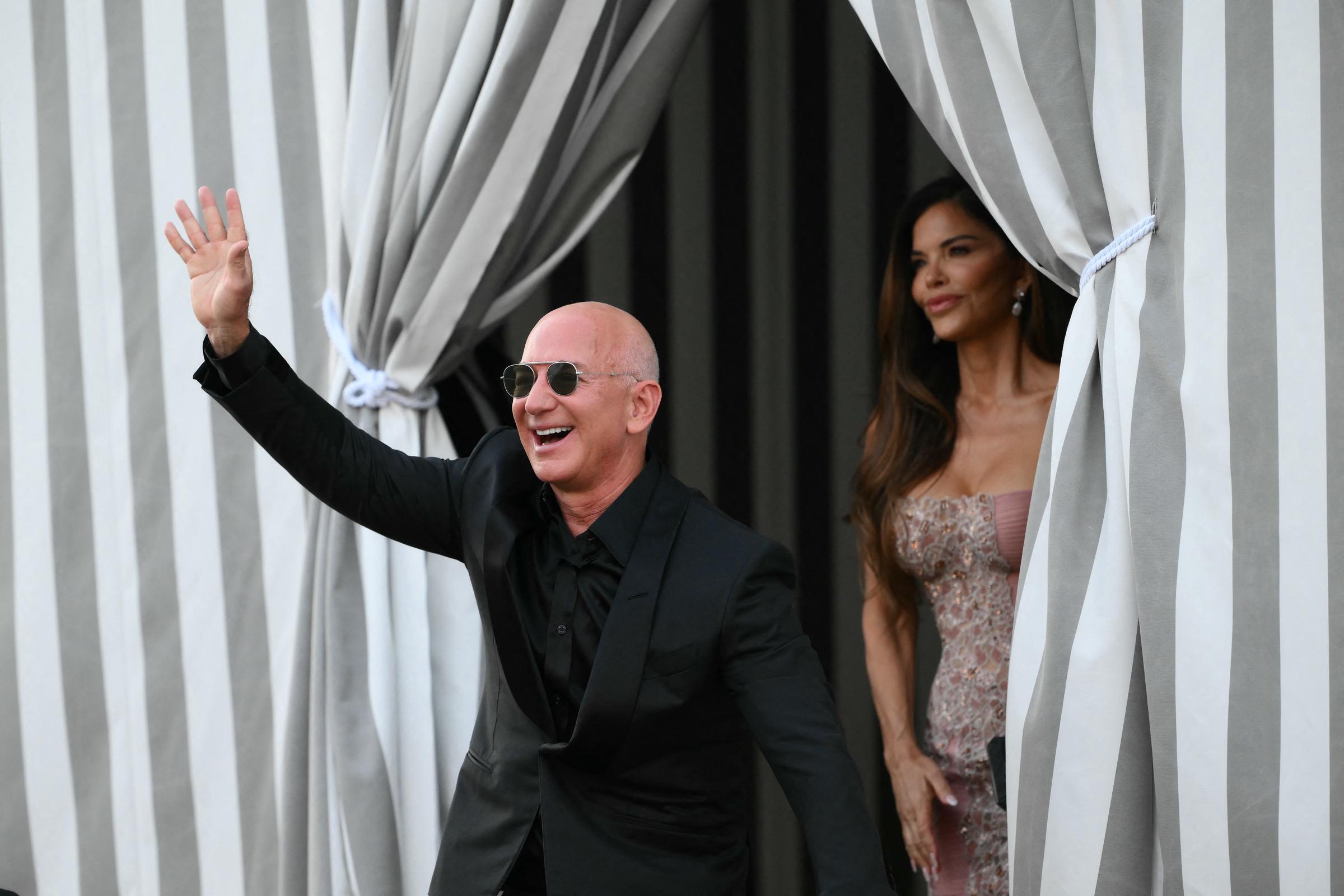 Jeff Bezos beams and waves energetically at onlookers, dressed in a sharp black suit and aviator sunglasses, while Lauren Sánchez stands gracefully behind him in a shimmering blush-toned gown with intricate beading, her long hair styled in loose waves as she gazes out from between elegant striped curtains.