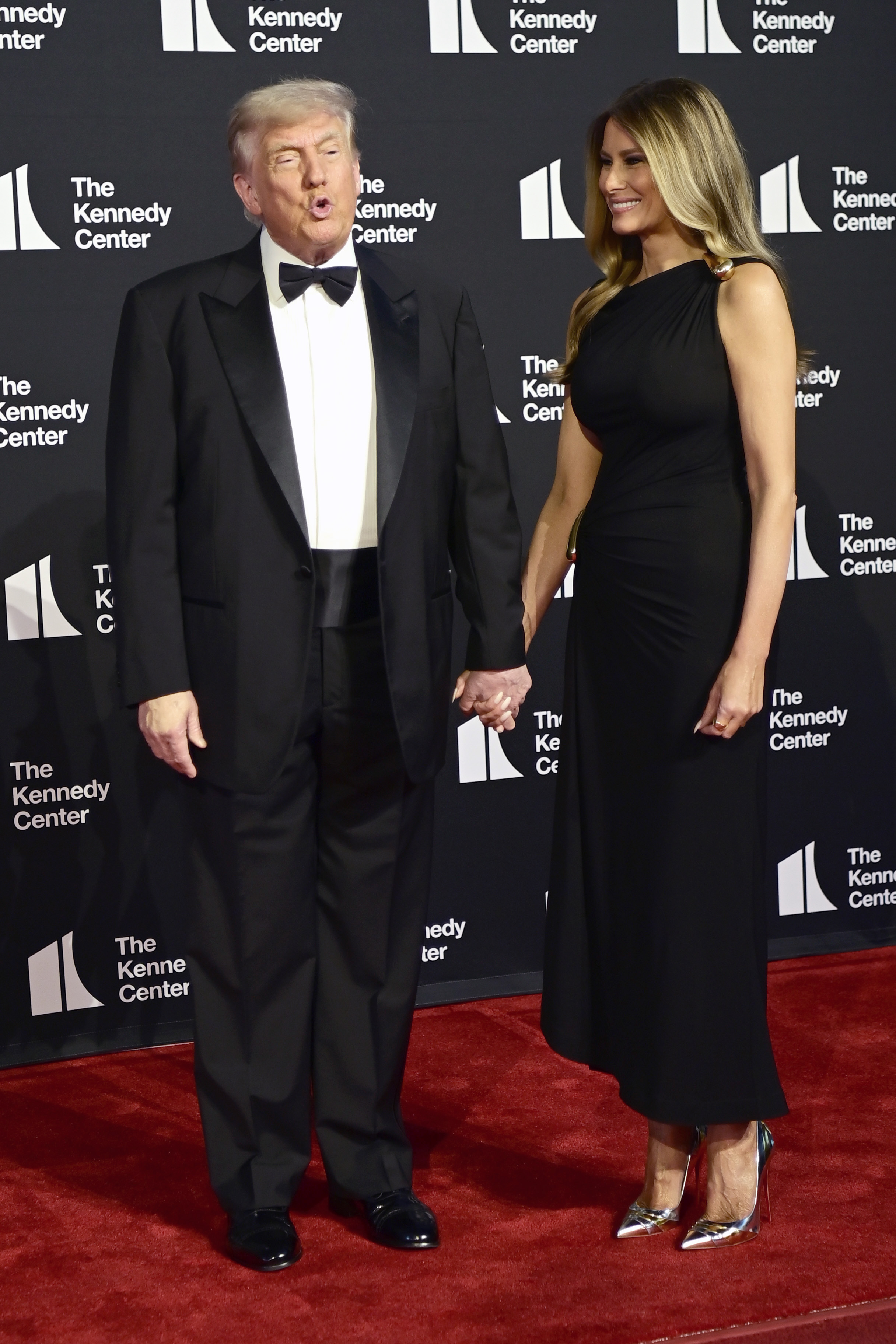 Donald and Melania hold hands on the red carpet, sharing a light moment. She smiles warmly in his direction, while he appears mid-sentence.